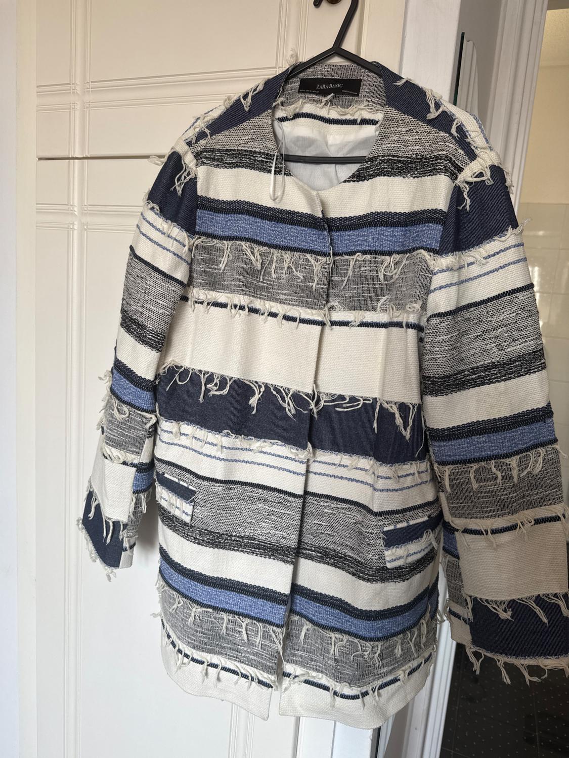 Coat hanging on a hanger, with horizontal stripes in shades of blue, white, and black, and frayed details. Visible label: ZARA BASIC.