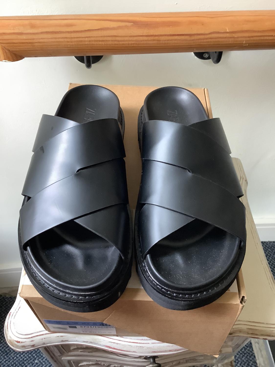 Black cross-strap sandals on a cardboard box under a wooden bar. Text: ZARA, 2731/120/800. Casual vibe.