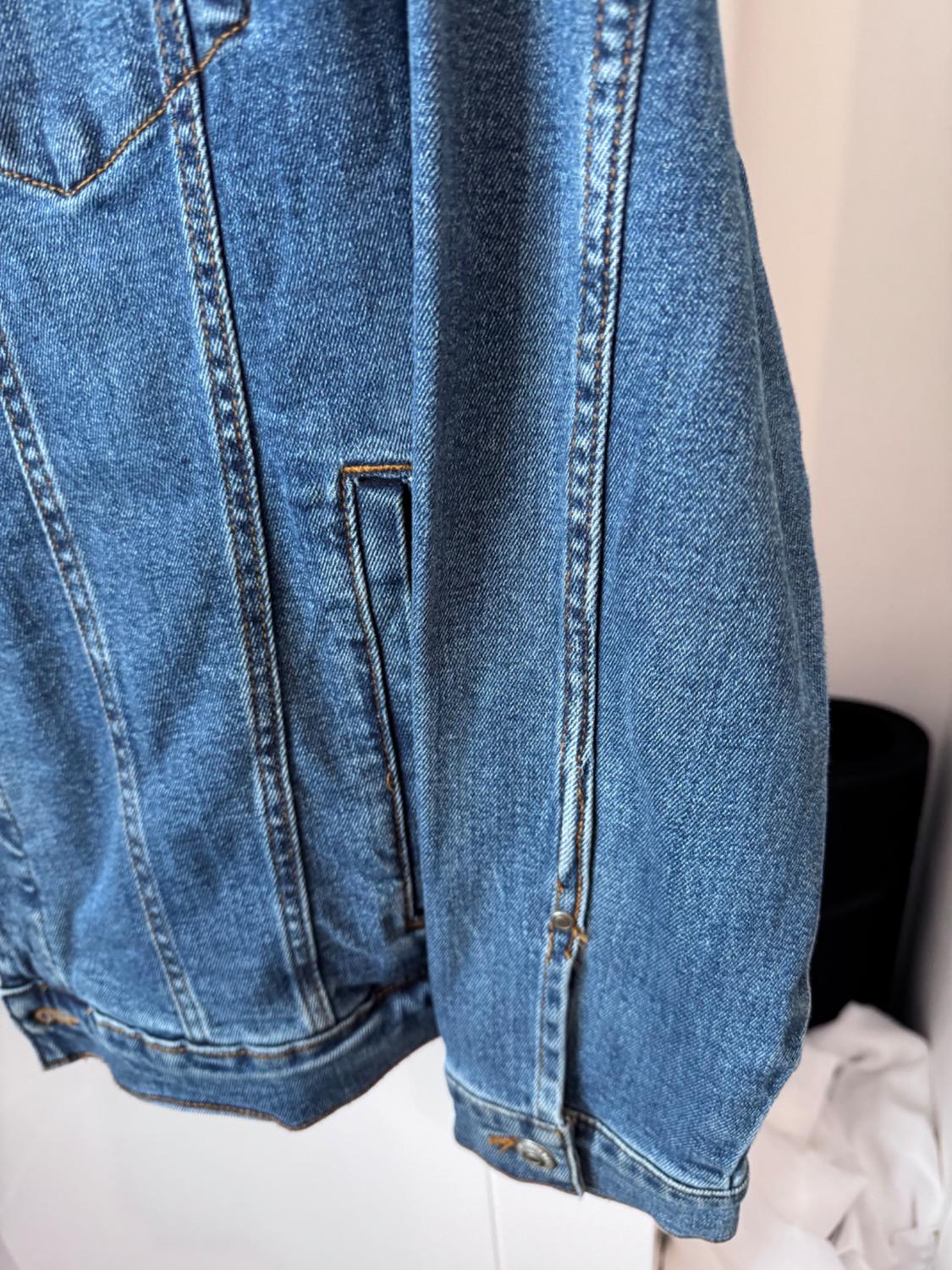 Blue denim jacket hanging, showing stitching details and side pocket. Black and white background.