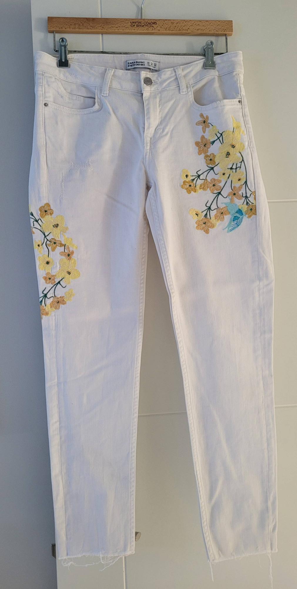 White trousers with yellow and blue floral embroidery, hanging on a wooden hanger from United Colors of Benetton.