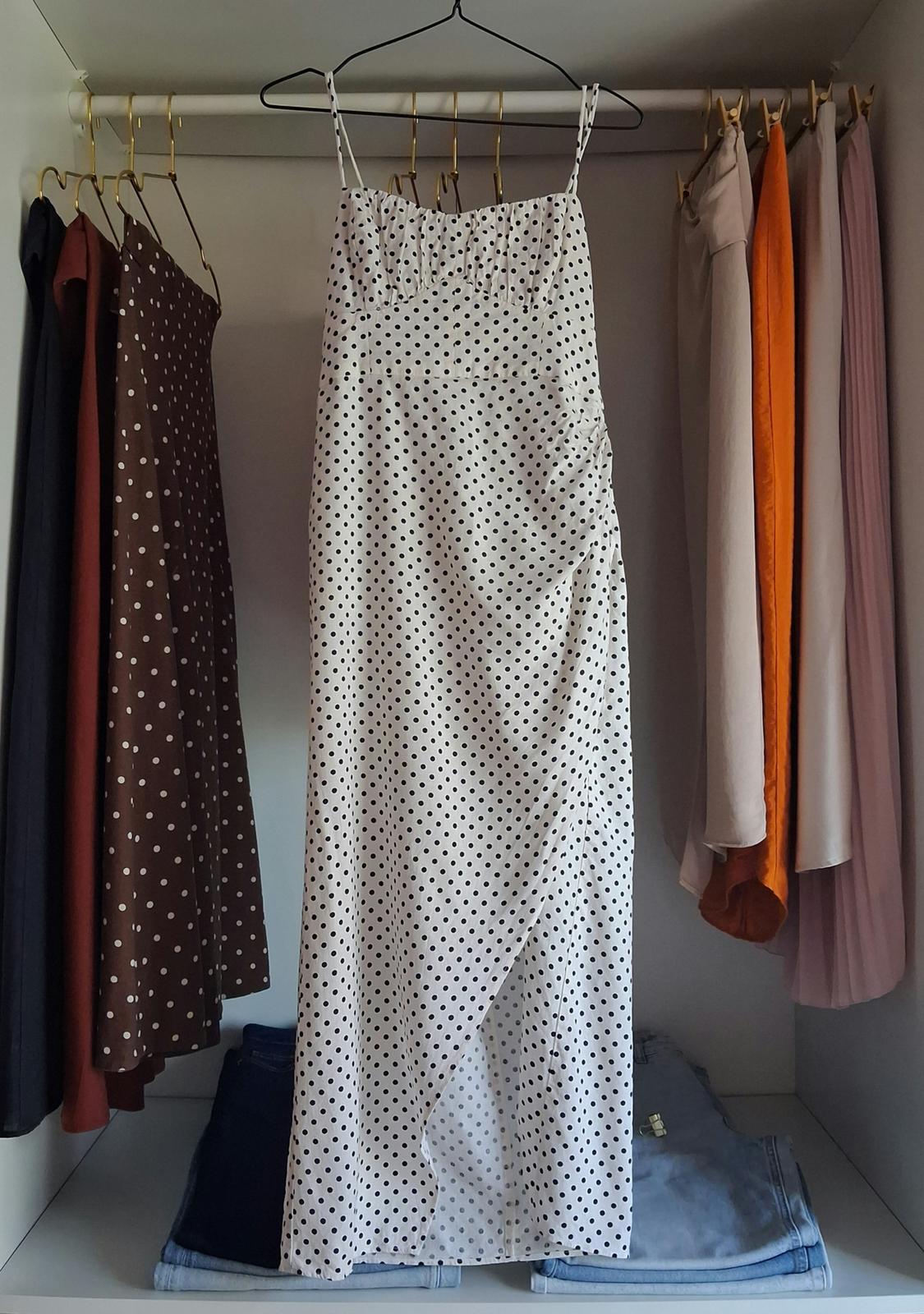 White dress with black polka dots and straps hanging in the centre of a wardrobe, surrounded by other garments and folded jeans at the bottom.