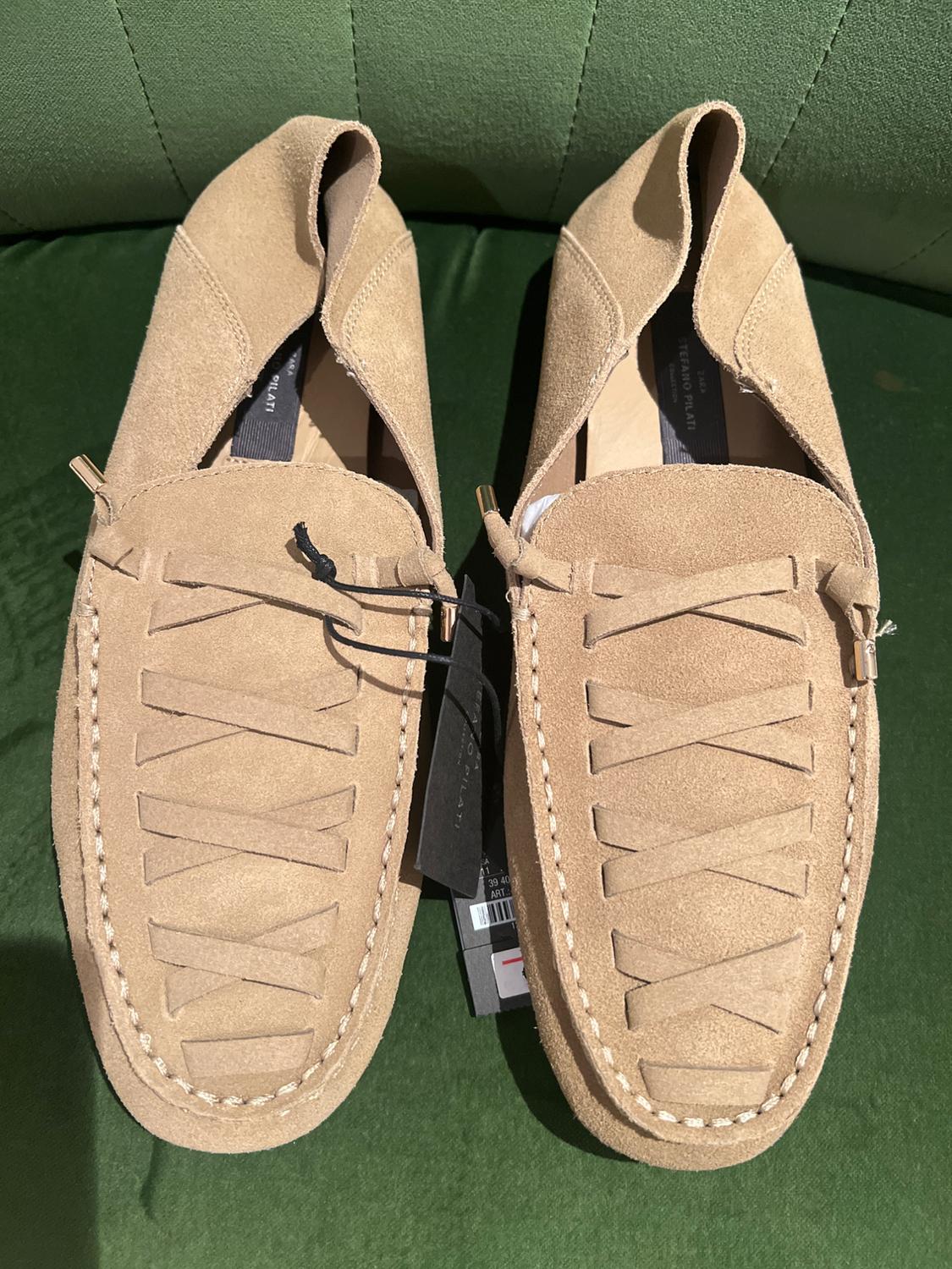 Pair of beige suede loafers with crossover straps on a green surface. Text: ZARA STEFANO PILATI.