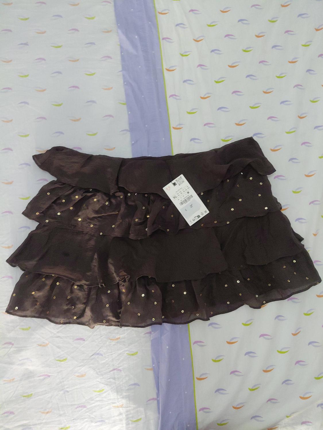 Dark brown short frilled skirt with gold sequins and a label, on a light-coloured sheet with a colourful patterned design.