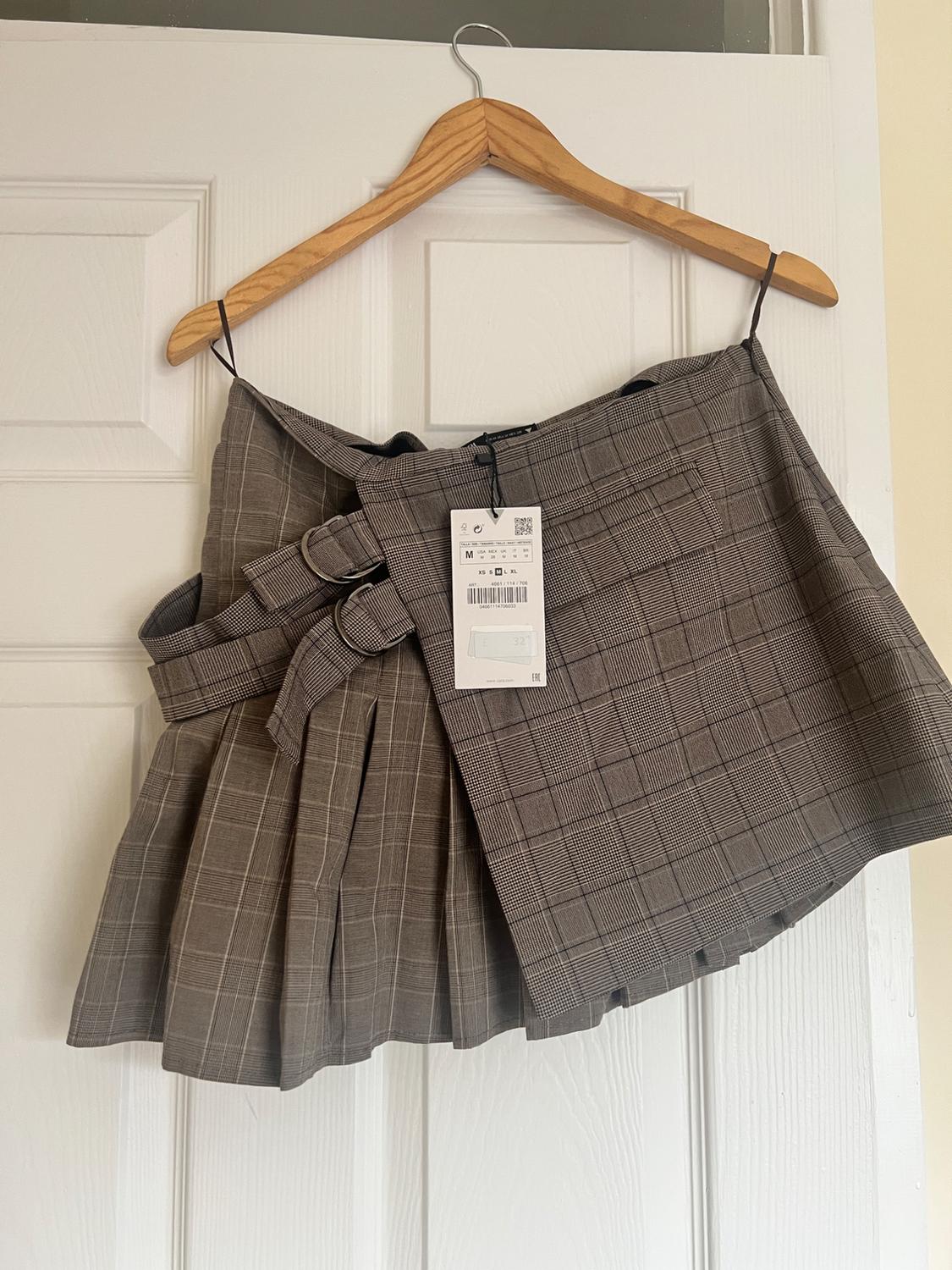 Brown checked skirt with belt and tag hanging on a wooden hanger over a white door.