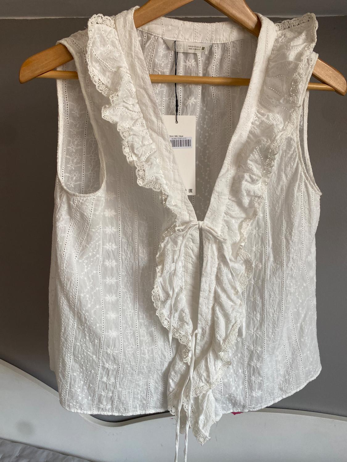 White sleeveless blouse with ruffles and lace, hanging on a wooden hanger. Label visible in the centre. Grey background.