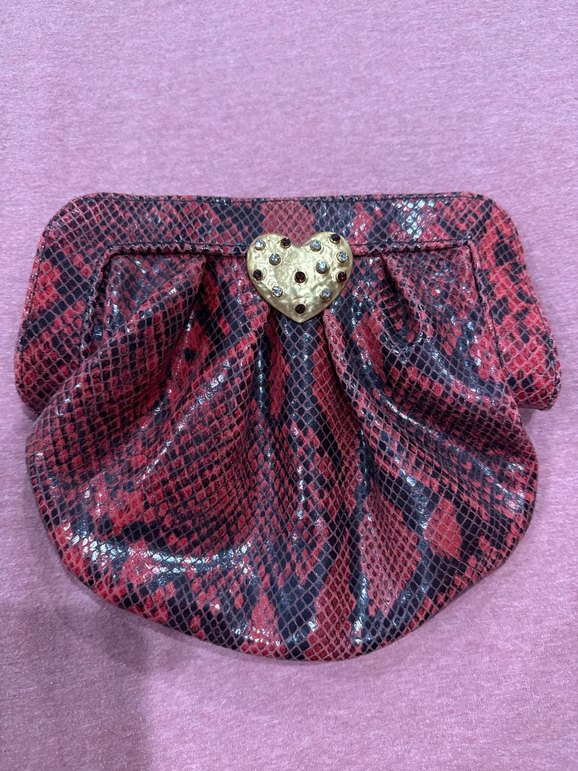 Handbag with snake print in shades of red and black, gold heart clasp with gemstones, on a pink background.