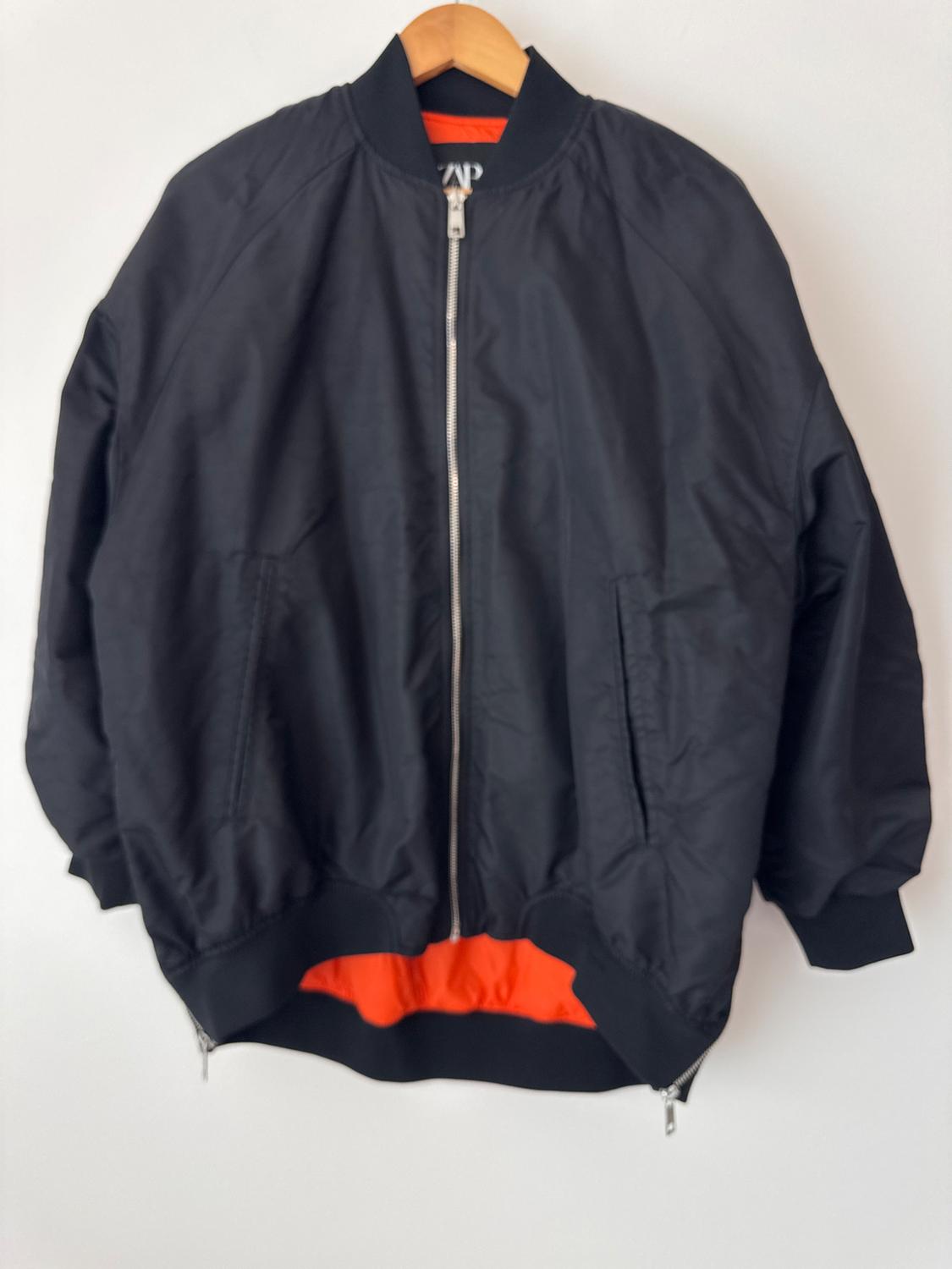 Black jacket with a front zipper and orange inner lining, hanging on a wooden hanger against a white background.