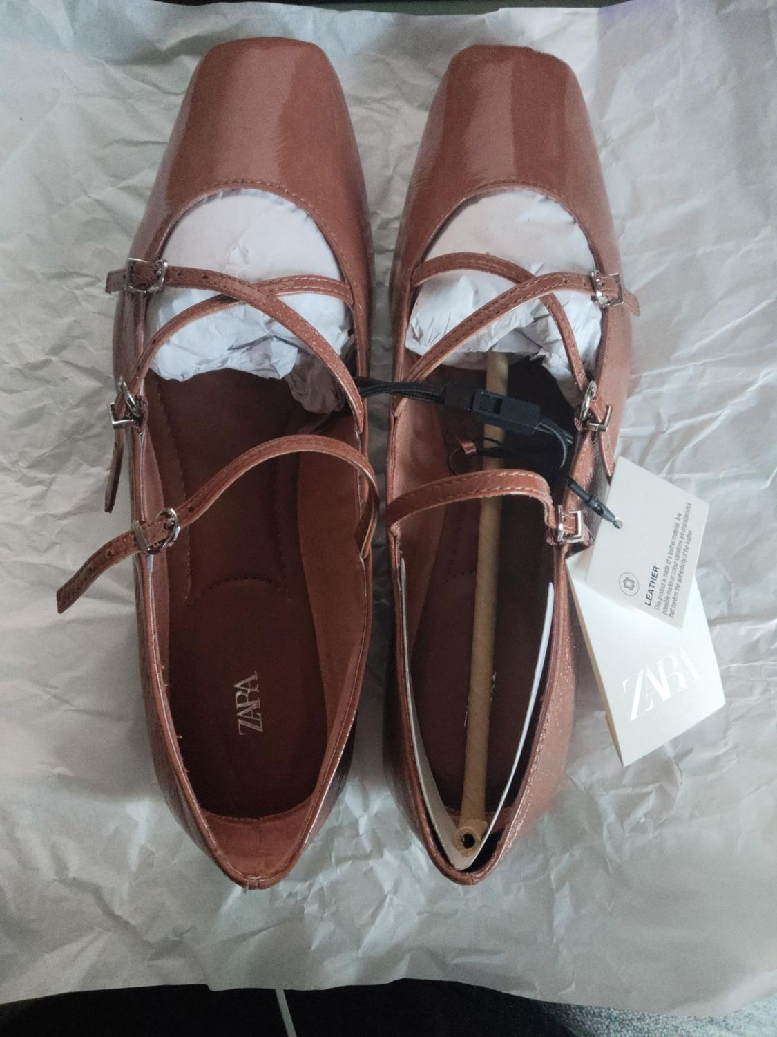 Brown patent leather shoes with crossover straps and buckles, on white paper. Visible label with Zara logo.