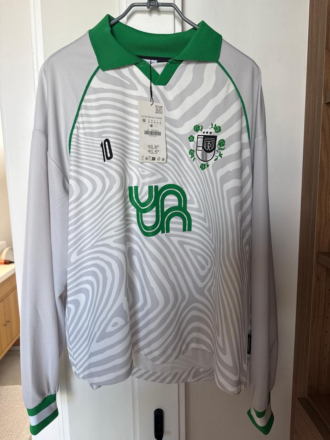 White t-shirt with grey wave print, green collar, number 10 and crest. Hanging tag with visible price.