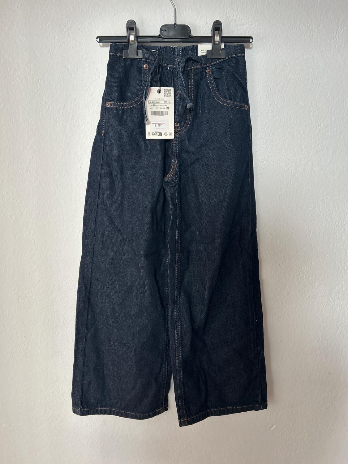 Dark denim jeans hung on a black hanger with visible tags, against a plain white background.