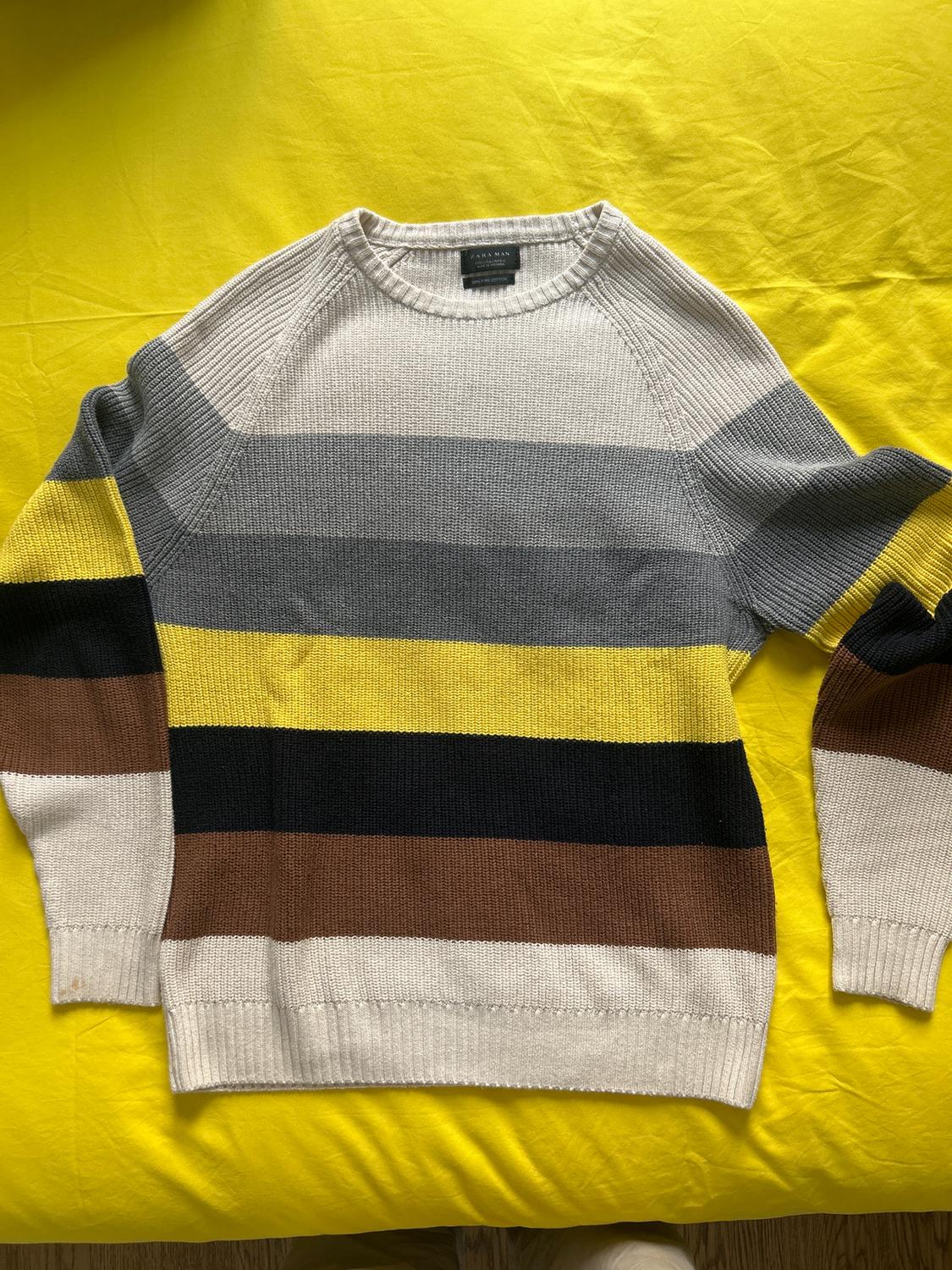 Horizontal striped jumper in shades of grey, yellow, black, and brown on a yellow surface. Label visible on the collar.