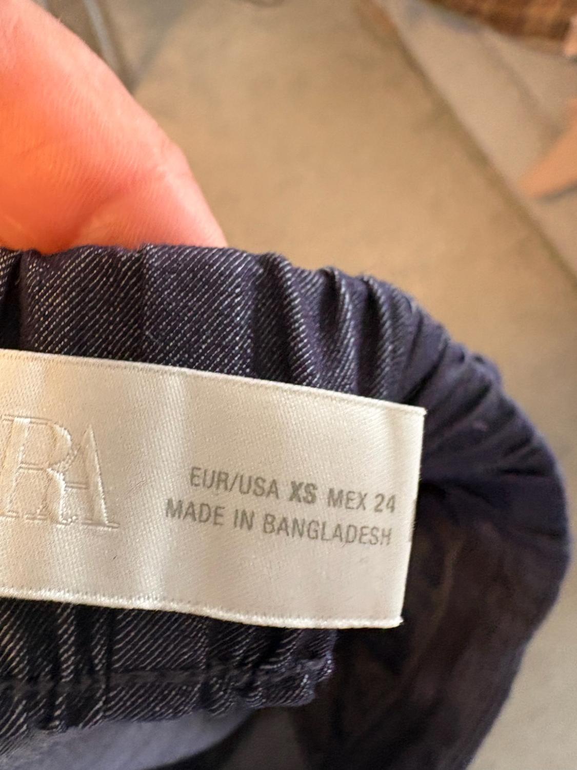 Close-up of fingers holding a blue elasticated waistband with a label that reads "EUR/USA XS MEX 24 MADE IN BANGLADESH".