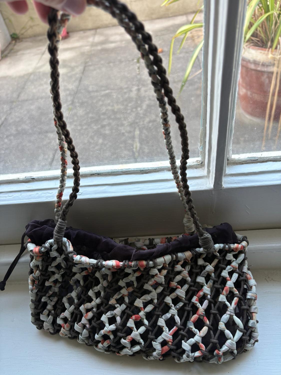 Hand holding a woven net bag with a dark lining and braided handles, resting on a white windowsill.
