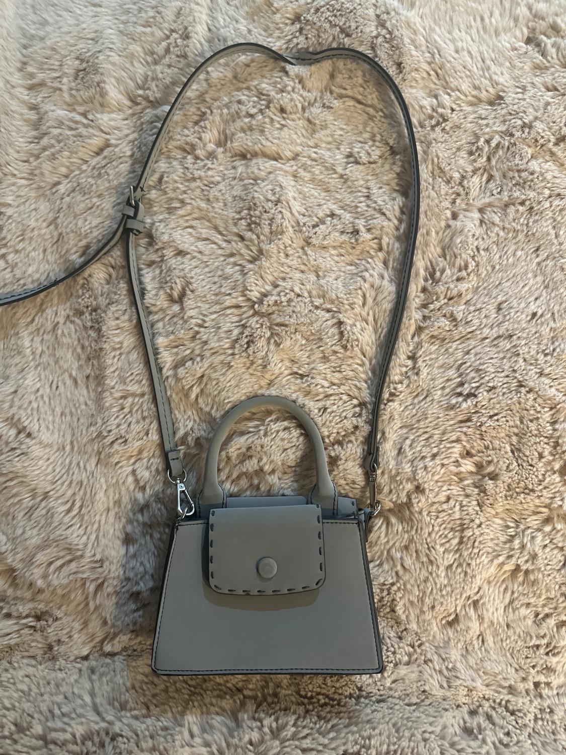 Small grey handbag with a top handle and a long adjustable strap on a beige shaggy rug.