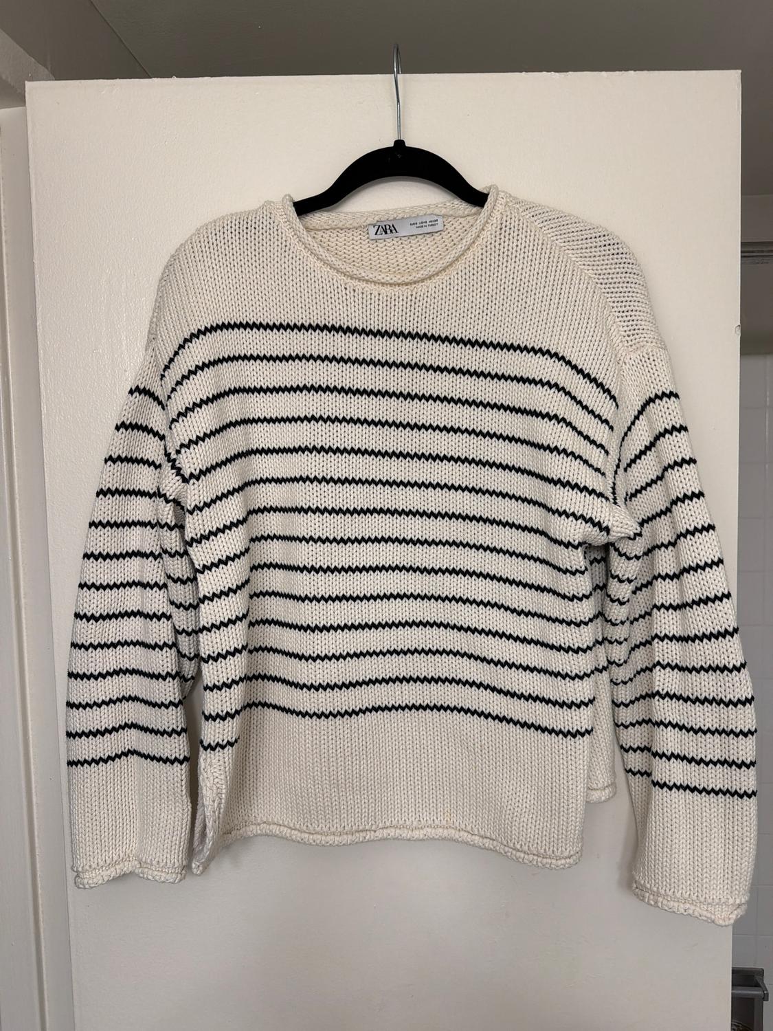 White knitted jumper with horizontal black stripes, hanging on a black hanger. Visible label: "ZARA". White background.