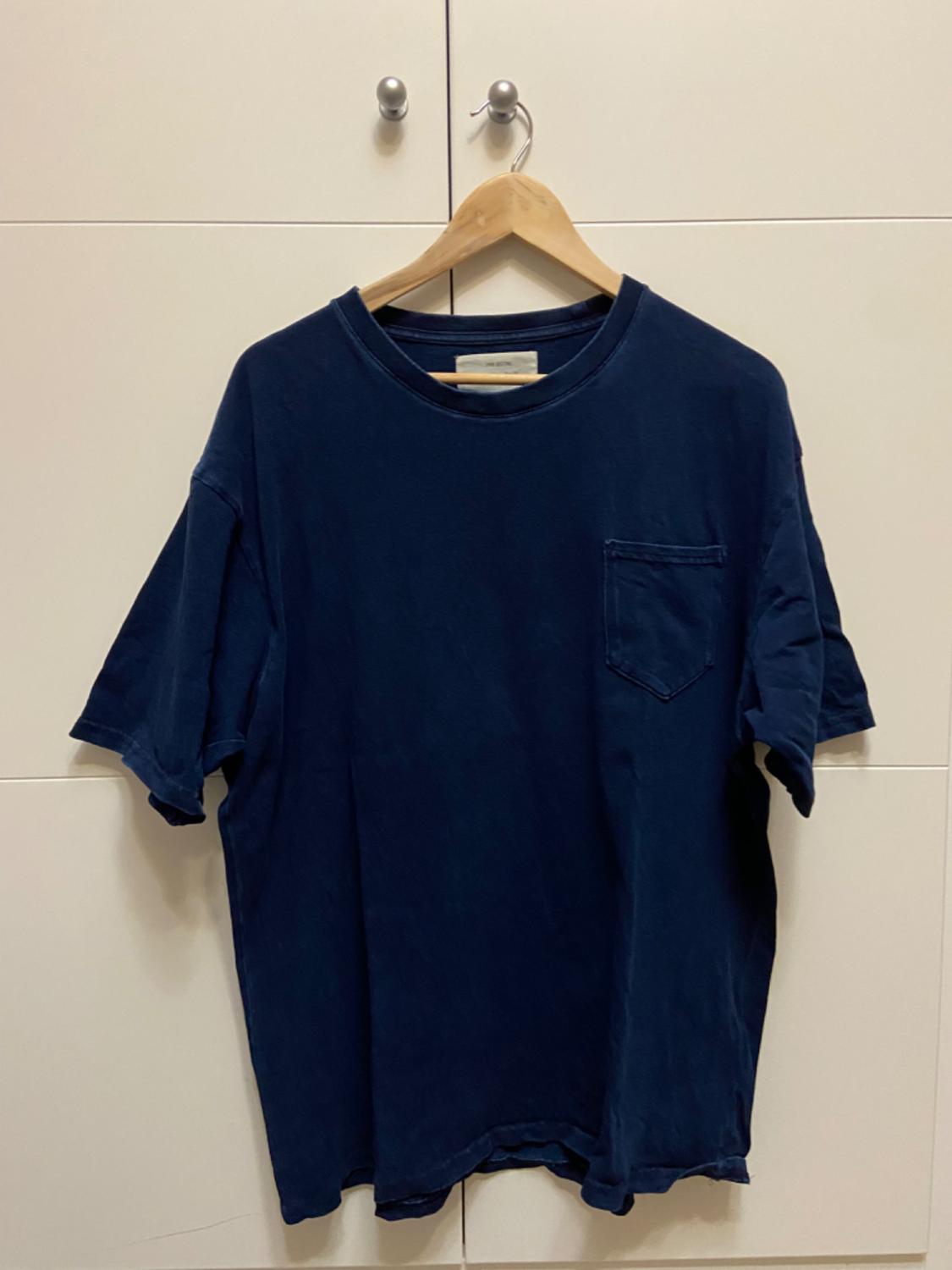 Dark blue t-shirt with a chest pocket, hanging on a wooden hanger in front of a white wardrobe with silver handles.