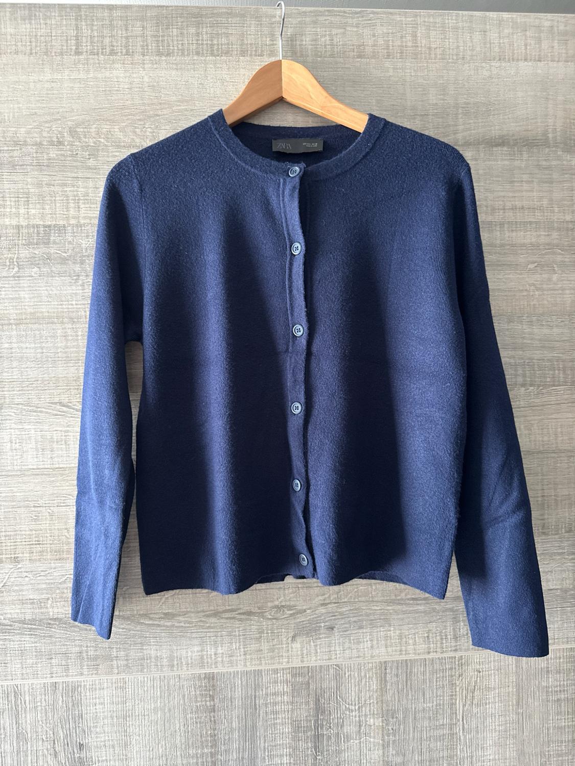 Navy blue knitted cardigan with round neck and buttons, hanging on a wooden hanger against a light wood background. Text: ZARA.