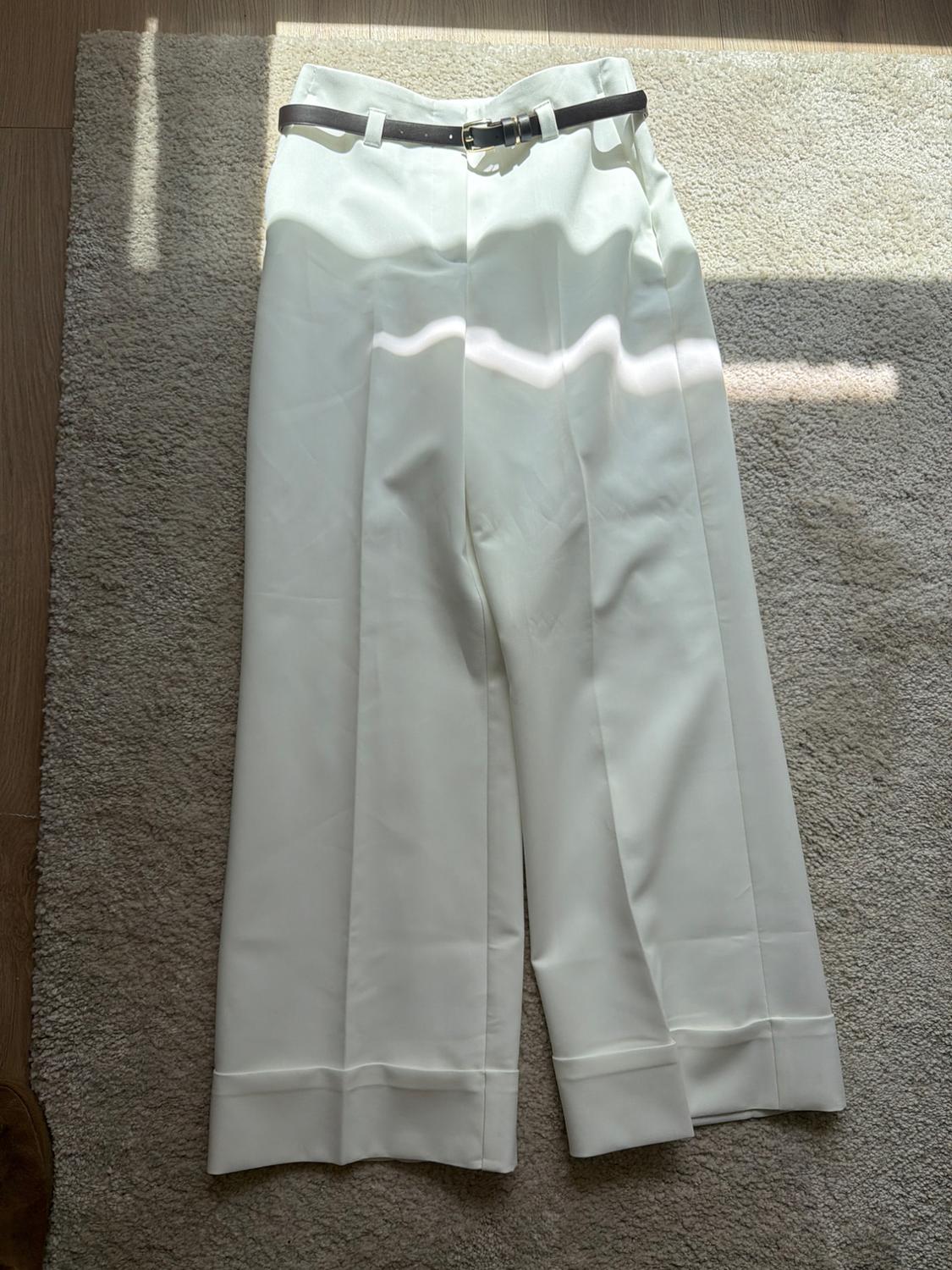 White wide-leg trousers with a black belt, placed on a beige rug illuminated by sunlight.