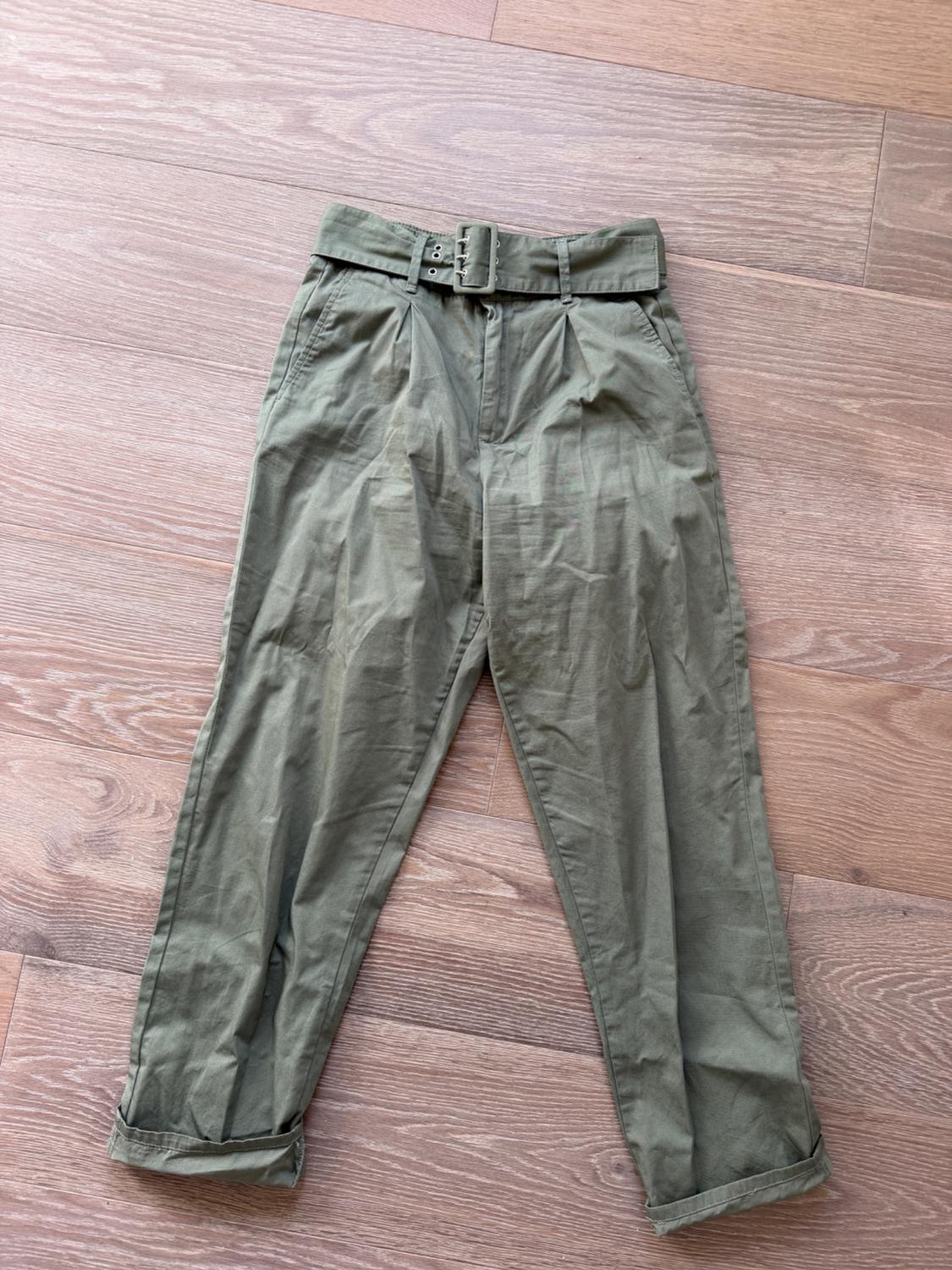 Green crinkled fabric trousers with a matching belt, placed on a light wooden floor.
