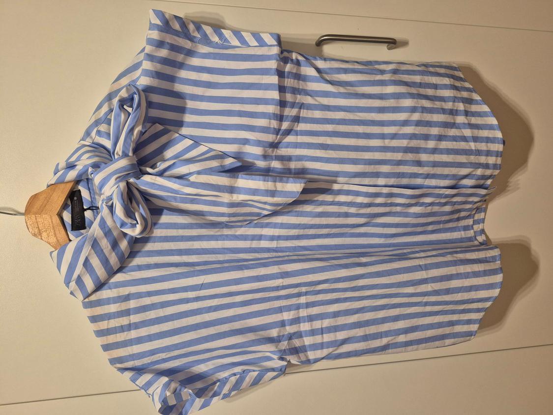 Short-sleeved shirt with blue and white stripes and a large bow at the neck, placed on a wooden hanger on a white surface.