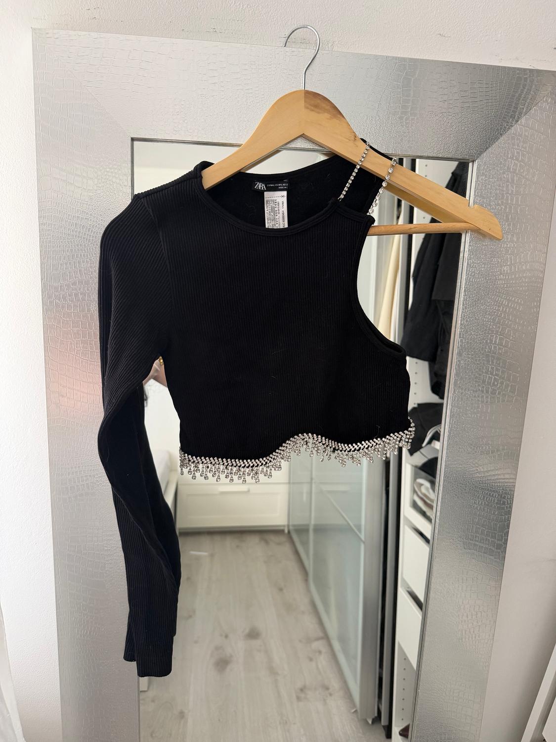 Black one-shoulder top with rhinestone detailing on the bottom edge, hanging on a hanger in front of a mirror.