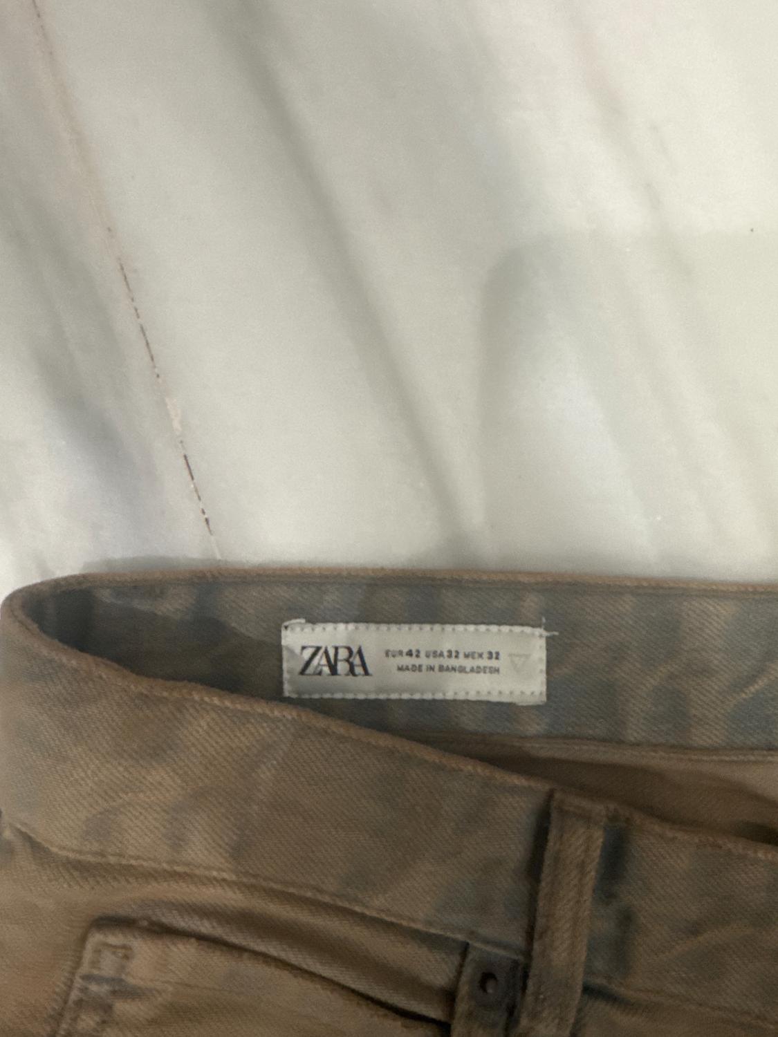 Λεπτομέρεια από τη μέση ενός καφέ παντελονιού πάνω σε λευκό ύφασμα. Ετικέτα με το κείμενο ZARA EUR 42 USA 32 MEX 32 MADE IN BANGLADESH.