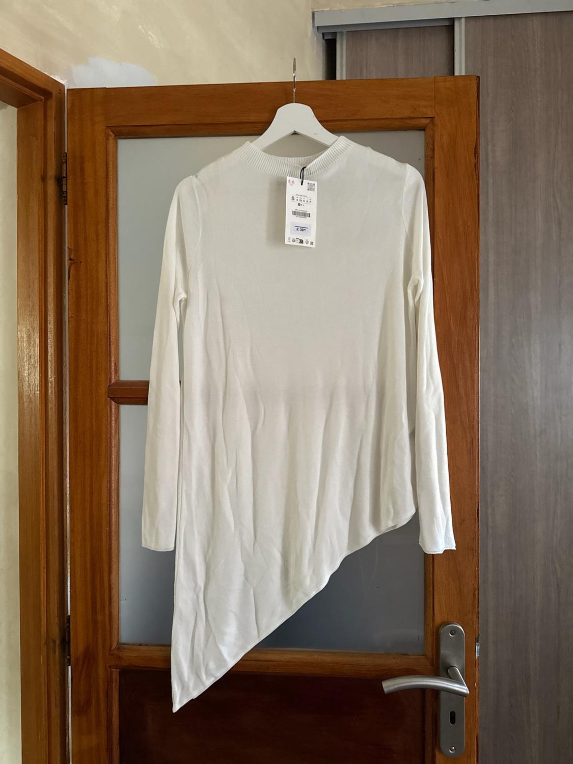 White long-sleeved top with an asymmetric hem and tag, hanging from a hanger on a wooden door with glass.