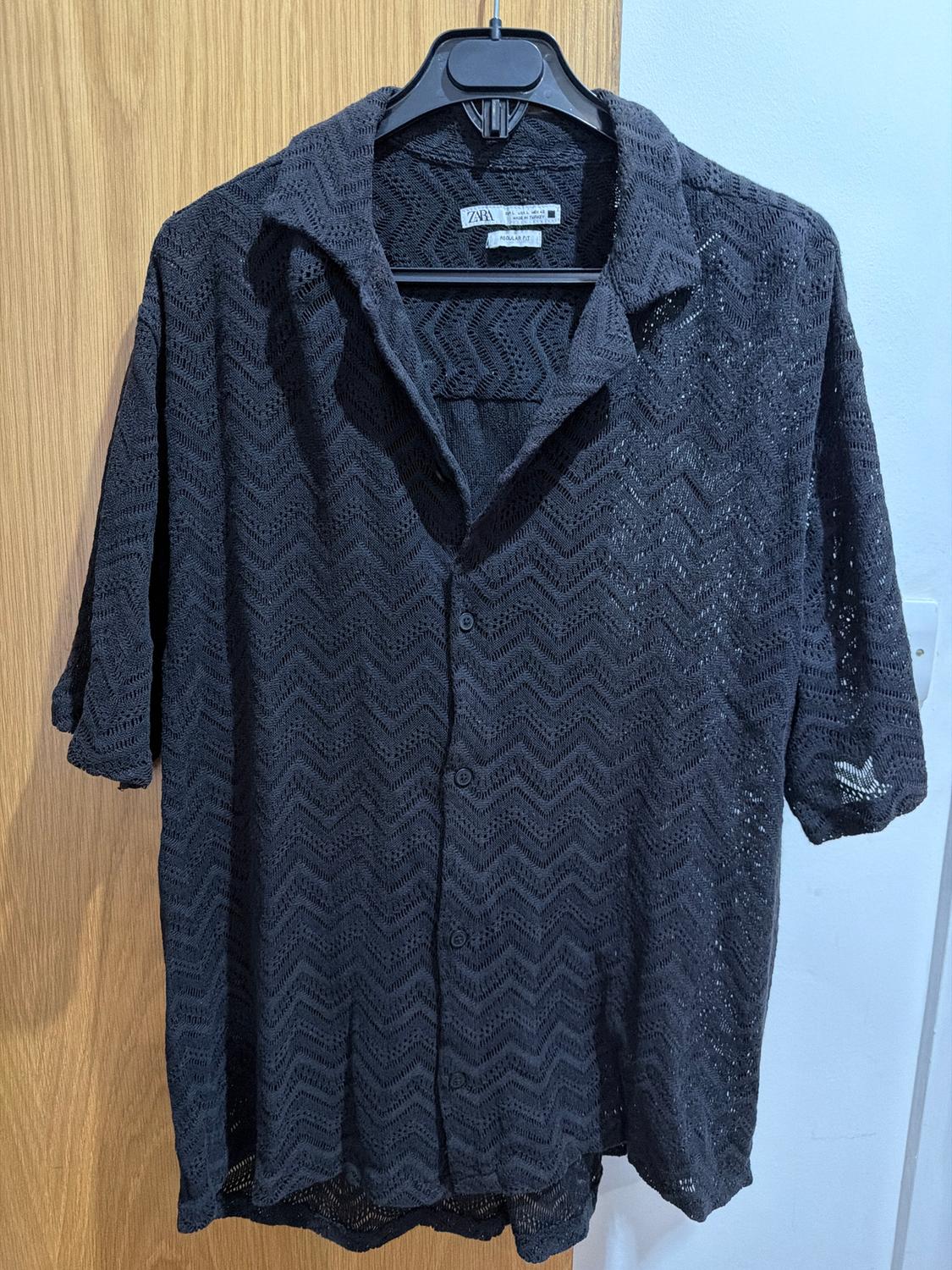 Black lace shirt with a zigzag pattern, short sleeves and a classic collar, hanging on a hanger over a wooden door.