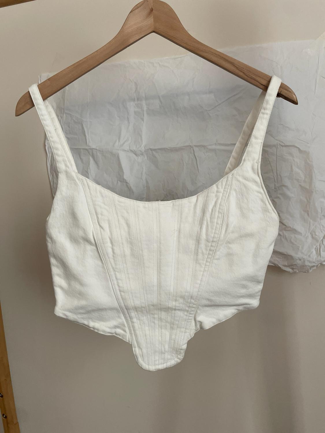 White corset-style top with vertical seams, hanging on a wooden hanger against a light background with crumpled white paper.
