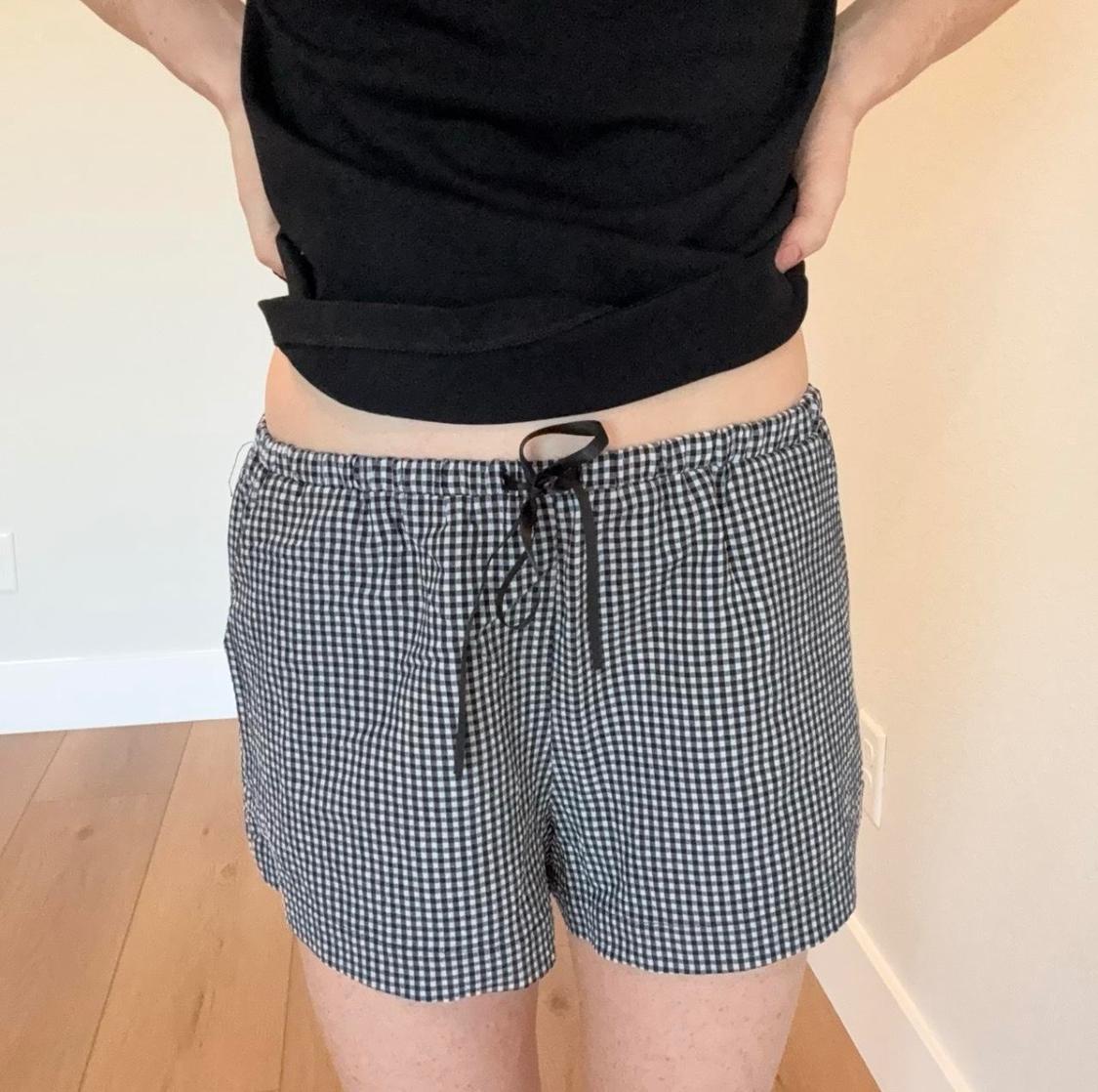 Person in a black t-shirt and black and white plaid shorts with a drawstring. Casual pose with hands on hips indoors.