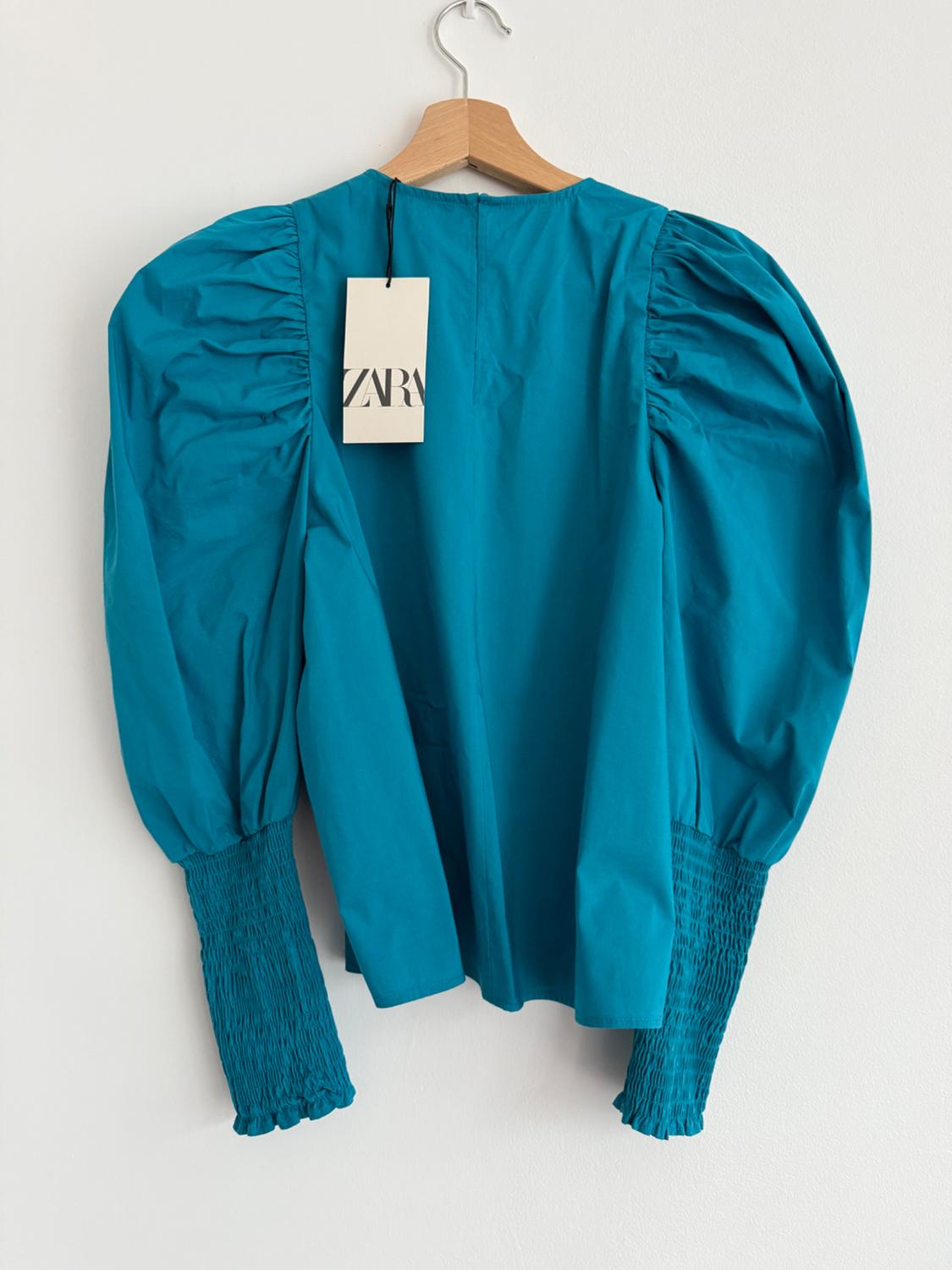 Blue blouse with puff sleeves and gathered cuffs, hanging on a wooden hanger. Label with text "ZARA".