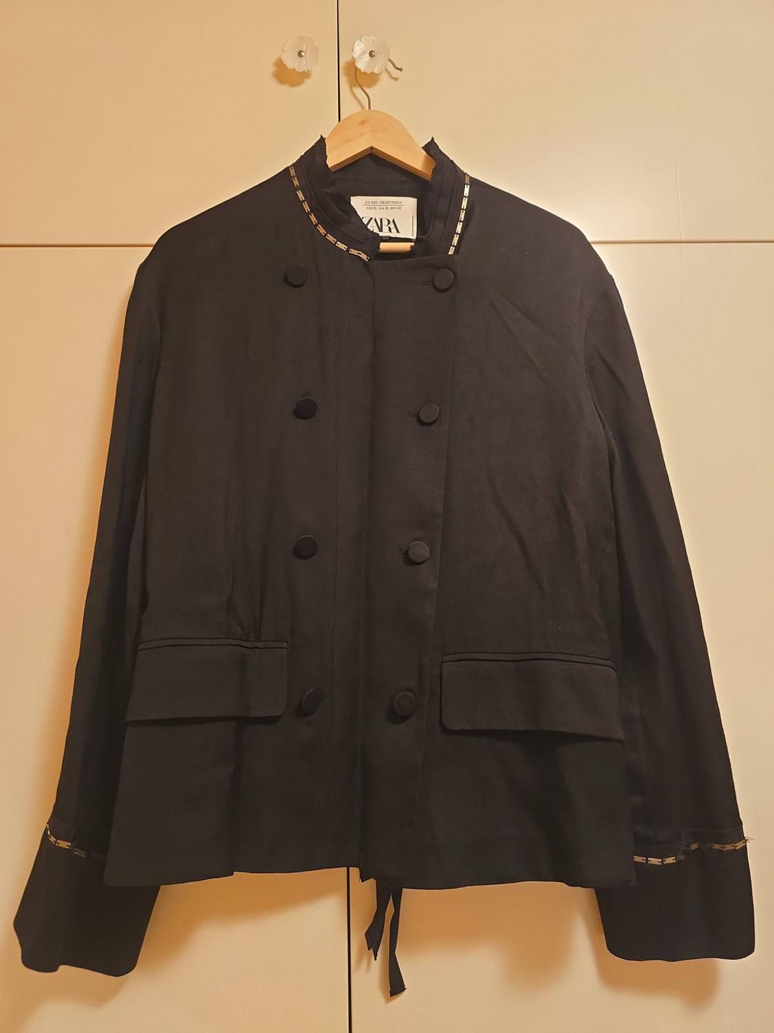 Black double-breasted jacket hanging on a hanger in front of a light-coloured wardrobe. It has detailing on the collar and cuffs, and a ZARA label.