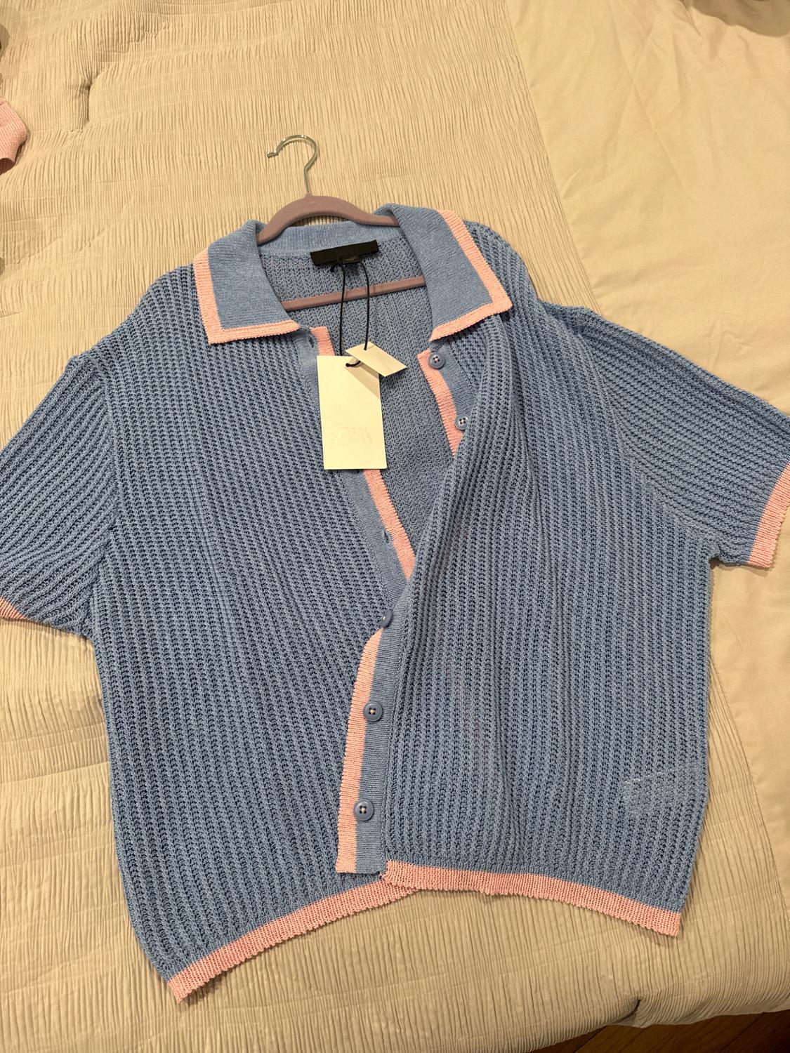 Blue cardigan with pink trim and buttons, hanging on a hanger over a beige bed. White tag visible.