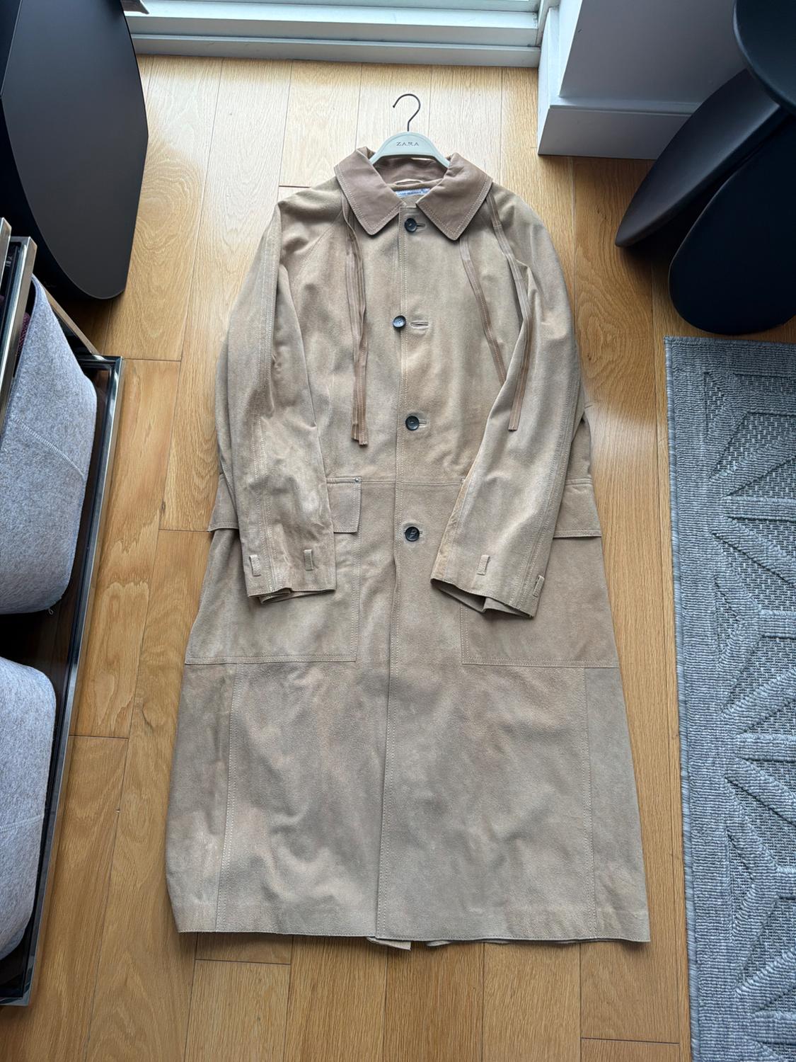 Beige suede coat with black buttons, hanging on a Zara hanger over a light wood floor next to dark furniture.