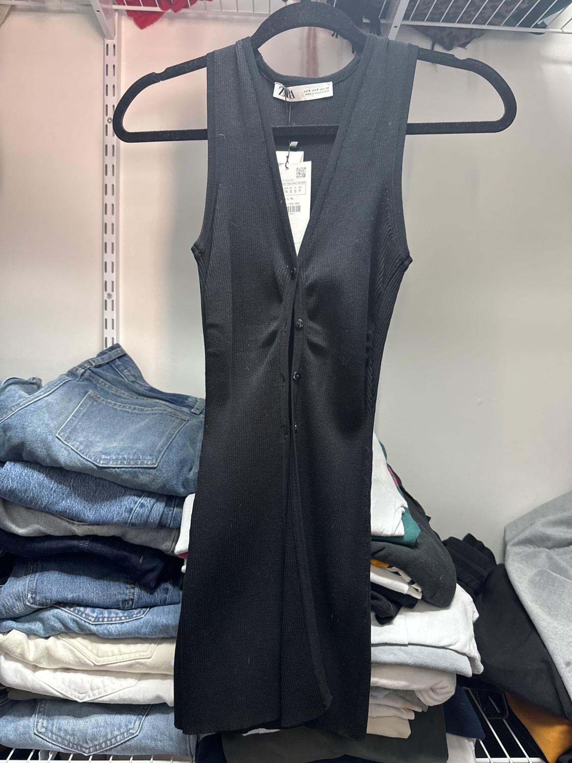 Sleeveless black dress with buttons down the front, hanging on a hanger. Background with shelves full of folded clothes, including jeans and t-shirts.