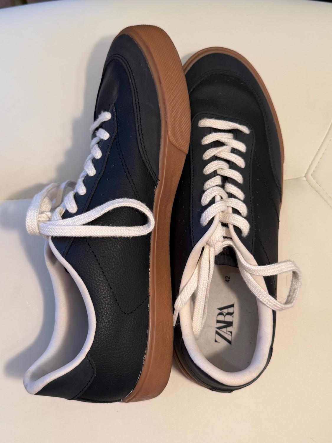 Black trainers with white laces and a brown sole on a white padded surface. The insole reads the text ZARA 42.