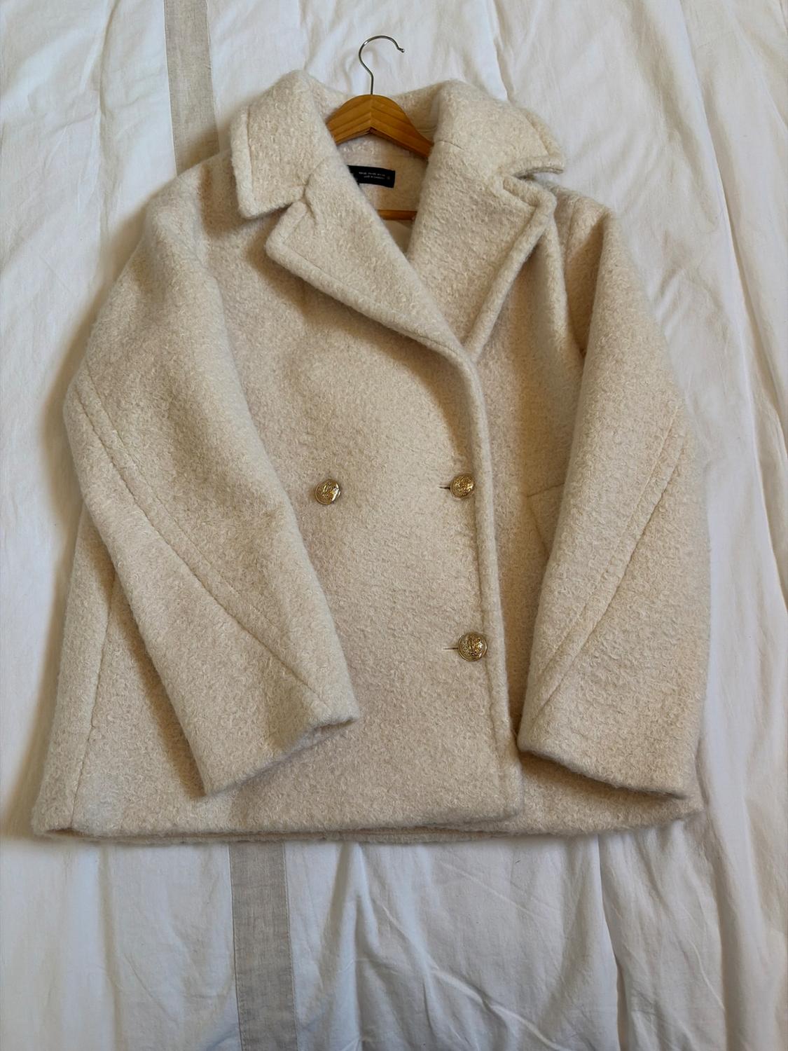 Cream-coloured textured double-breasted coat with gold buttons, on a wooden hanger against a crumpled white sheet. Relaxed style.
