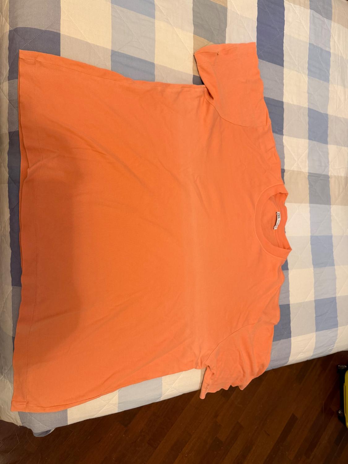 Orange extended short-sleeved T-shirt laid out on a bed with a blue and beige checked bedspread.