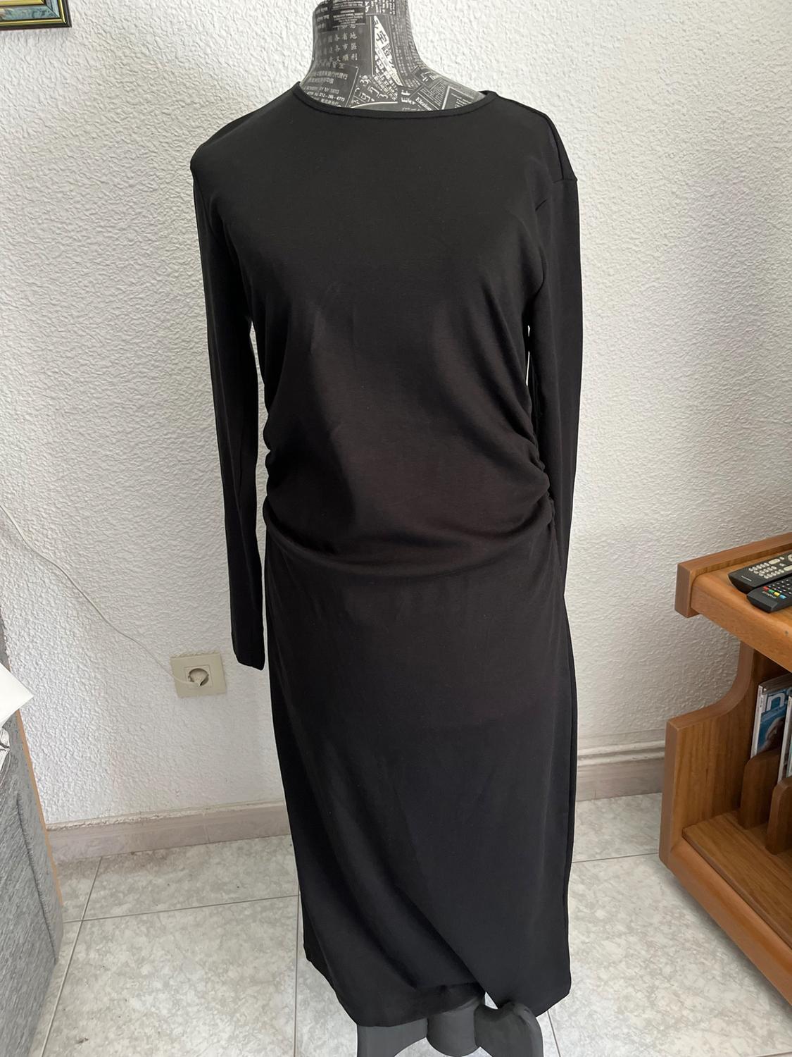 Long-sleeved black gathered dress on a newspaper-print mannequin in a room with a white wall and tiled floor.
