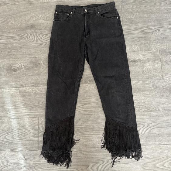 Black denim trousers with long fringing at the hems, placed on a light grey wooden floor.