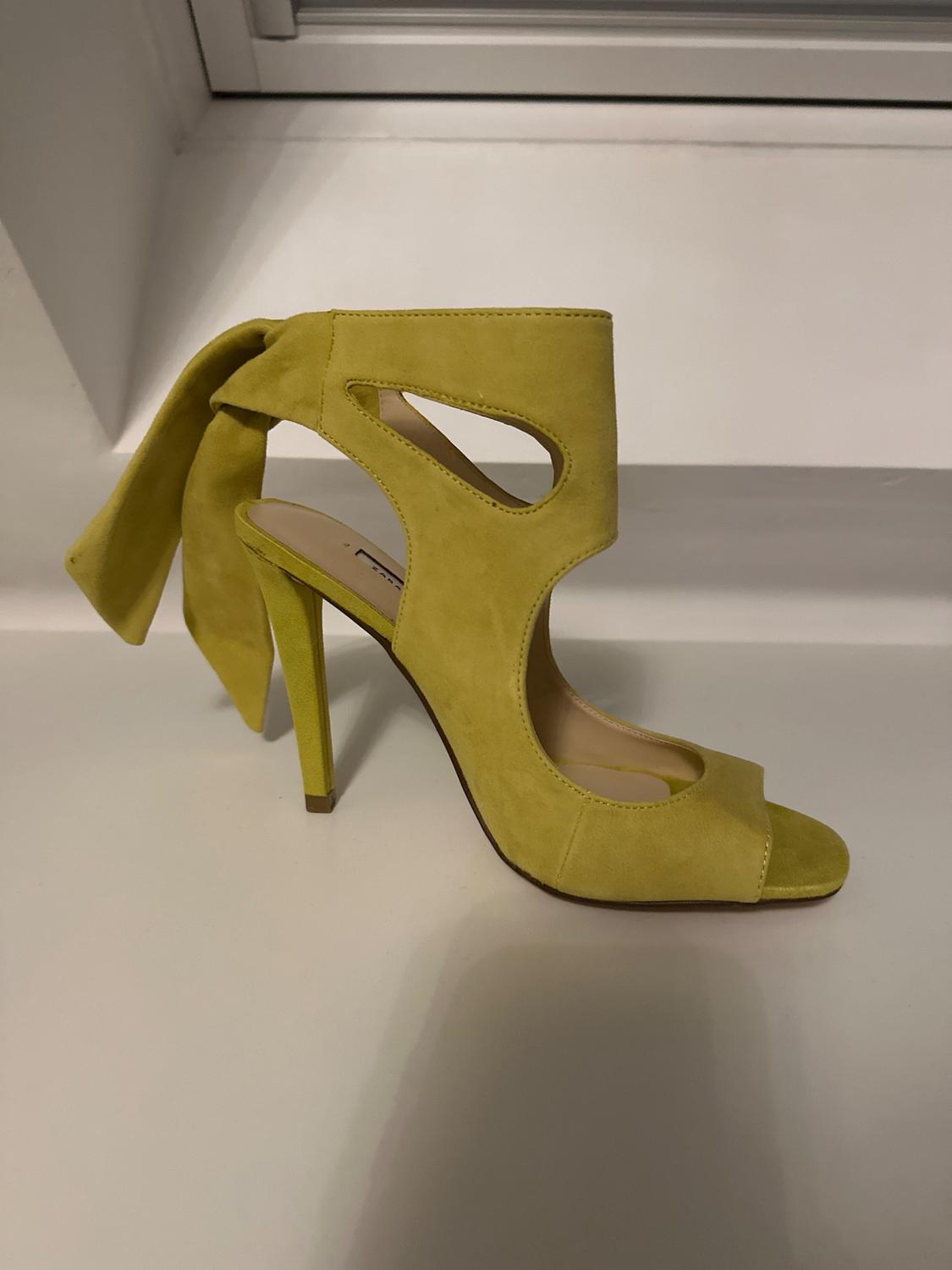 Yellow high-heeled shoe with side cut-outs and a bow on the heel, on a white surface in front of a window.