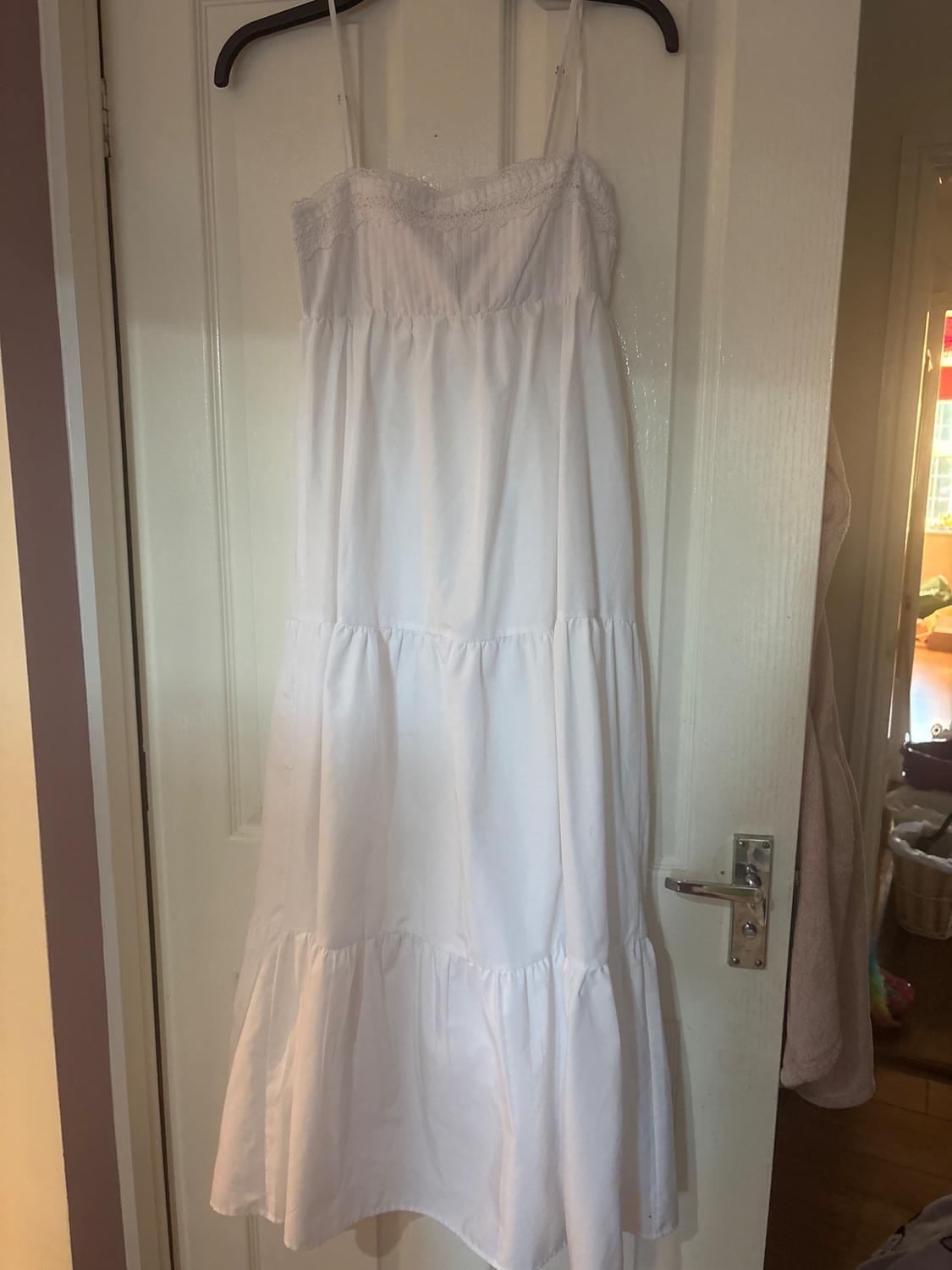 White strappy maxi dress with lace detailing and a tiered skirt, hanging on a white door in a relaxed, homely setting.