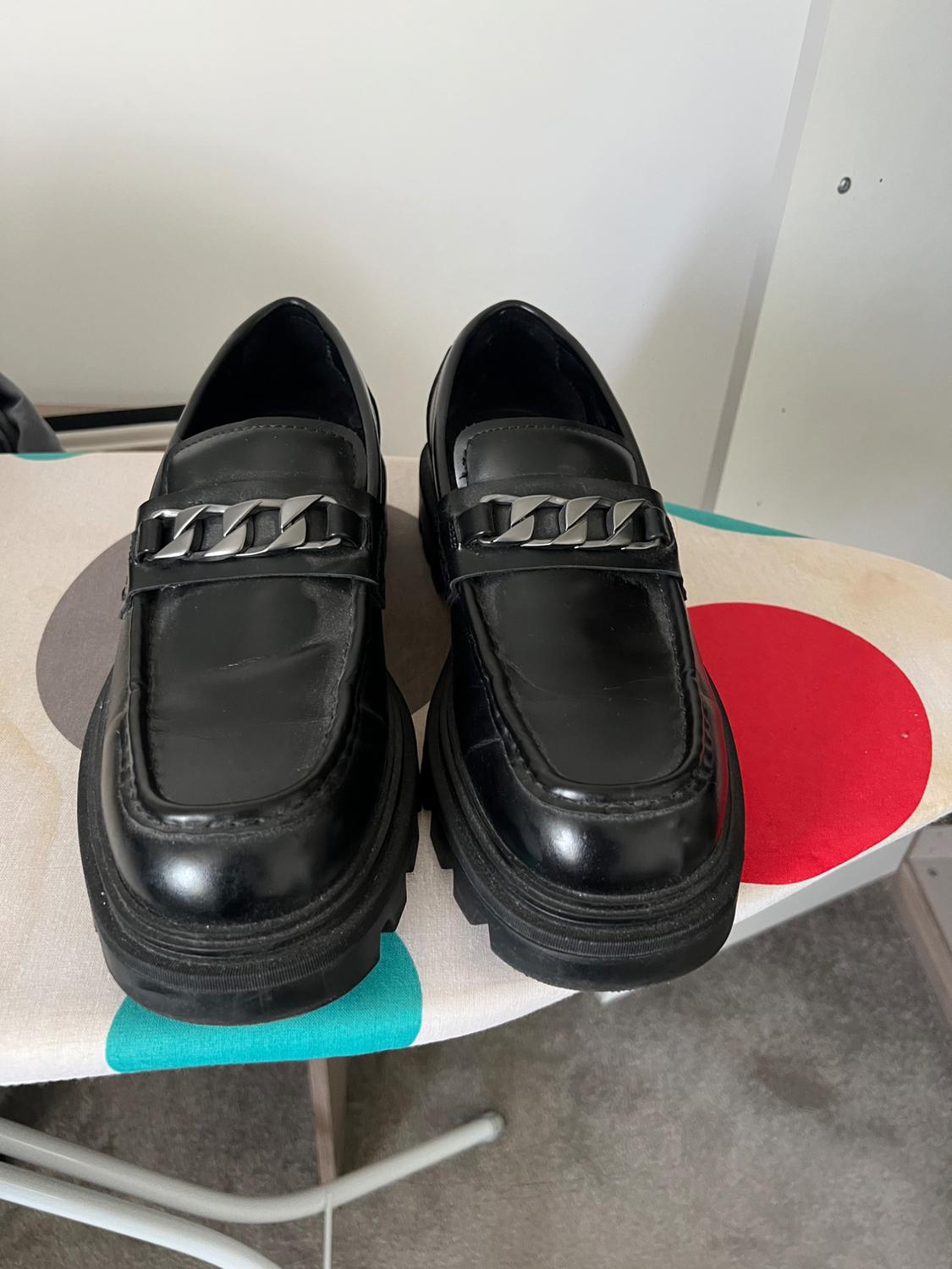 Black loafers with a chunky sole and metallic chain detail, resting on an ironing board with colourful circles.