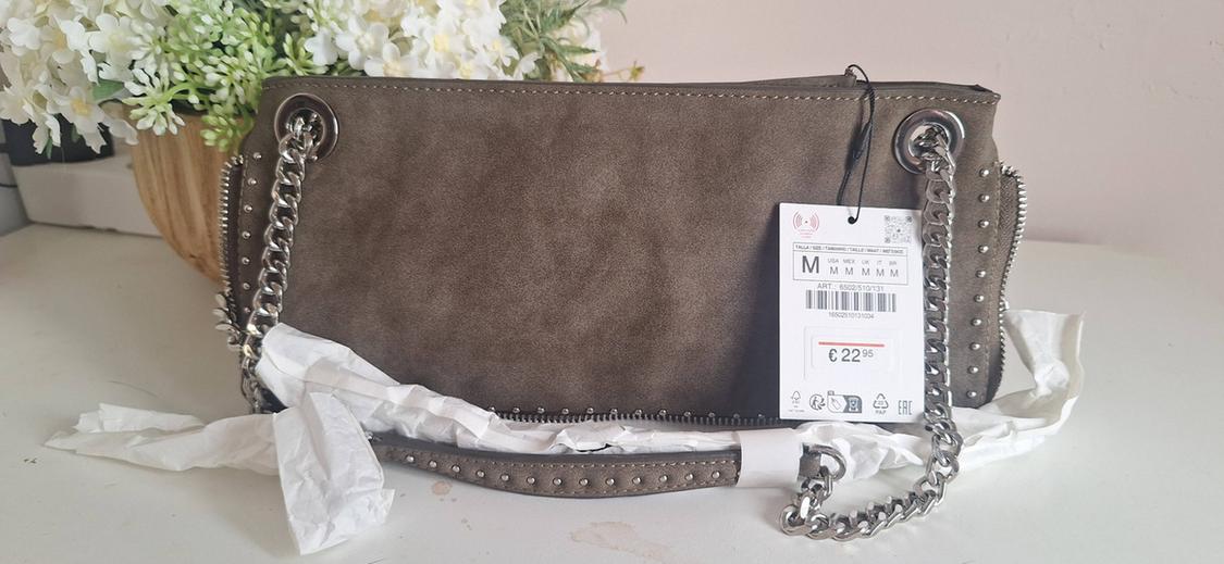 Brown bag with silver-coloured chain and studs on a white table with flowers. Label with text: M, € 22.95.
