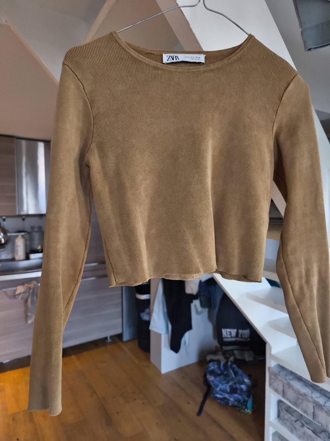 Brown long-sleeved jumper hanging on a hanger in a modern interior. Visible label with the text "Zara". Background with kitchen and staircase.