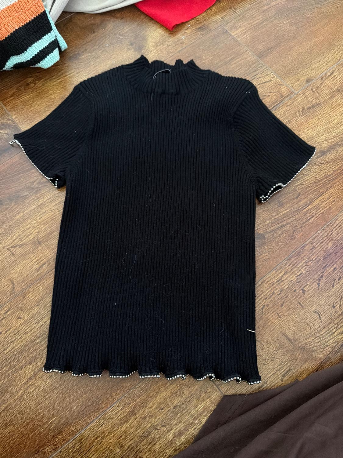 Black ribbed knit t-shirt with short sleeves and pearl details on the edges, on a wooden floor.