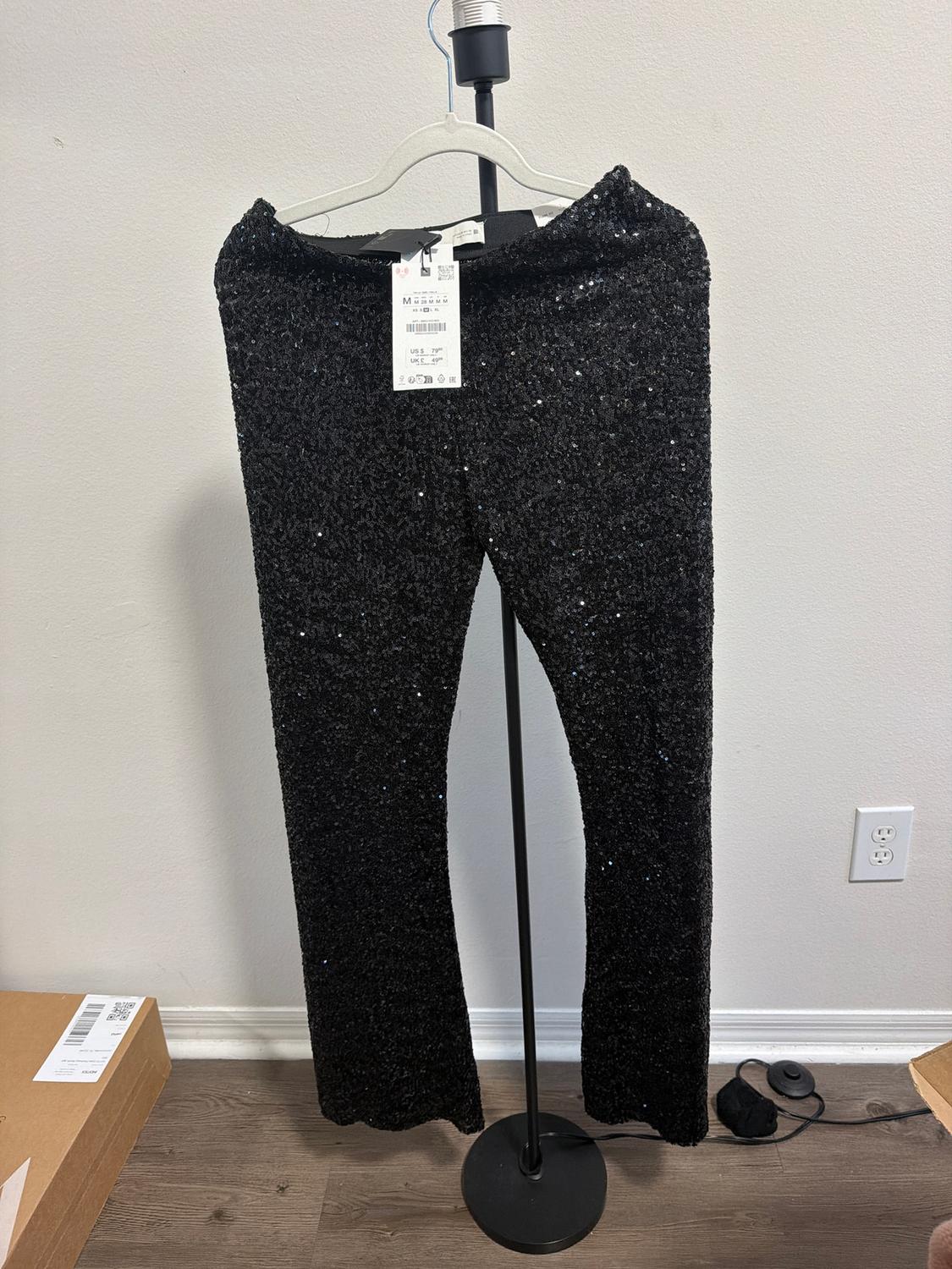 Black sequined pants hanging on a black clothes rack. Visible tag with product details. White background and wooden floor.