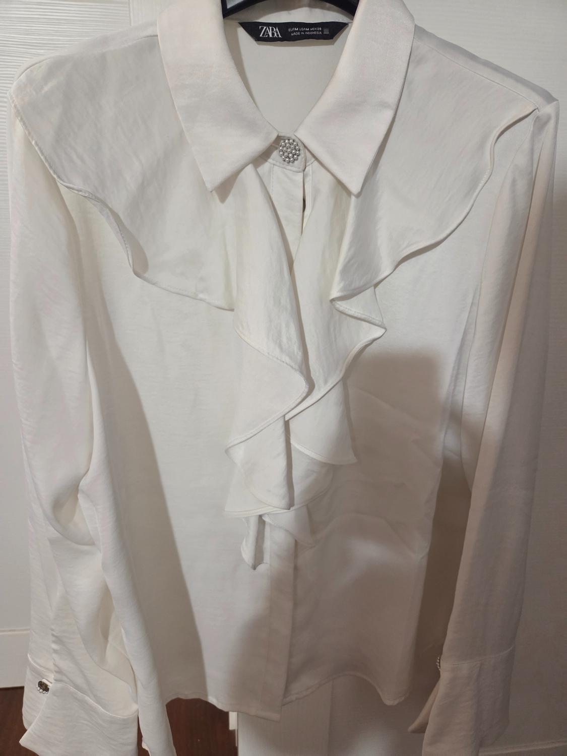 White blouse with frills and classic collar, hanging on a hanger. Decorative button detail on the collar. Visible label: ZARA.
