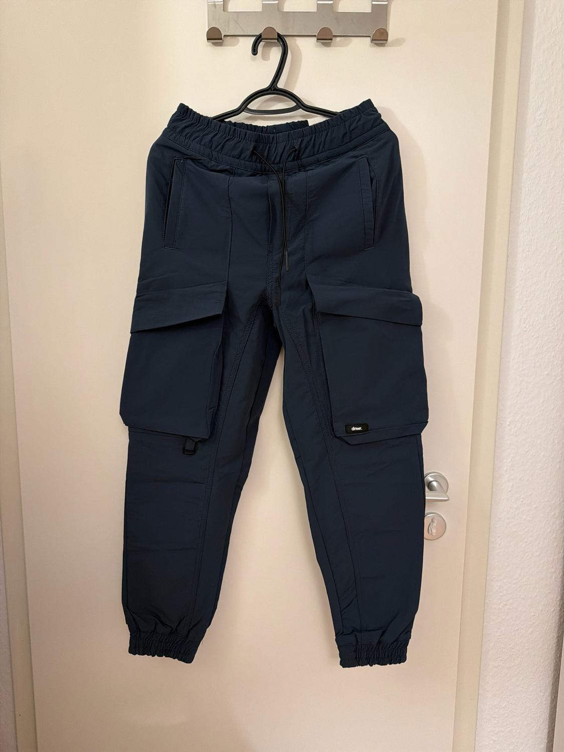 Navy blue cargo trousers hanging on a white door. Casual style with an elasticated waist and ankles, side pockets, and "dnwr." text.