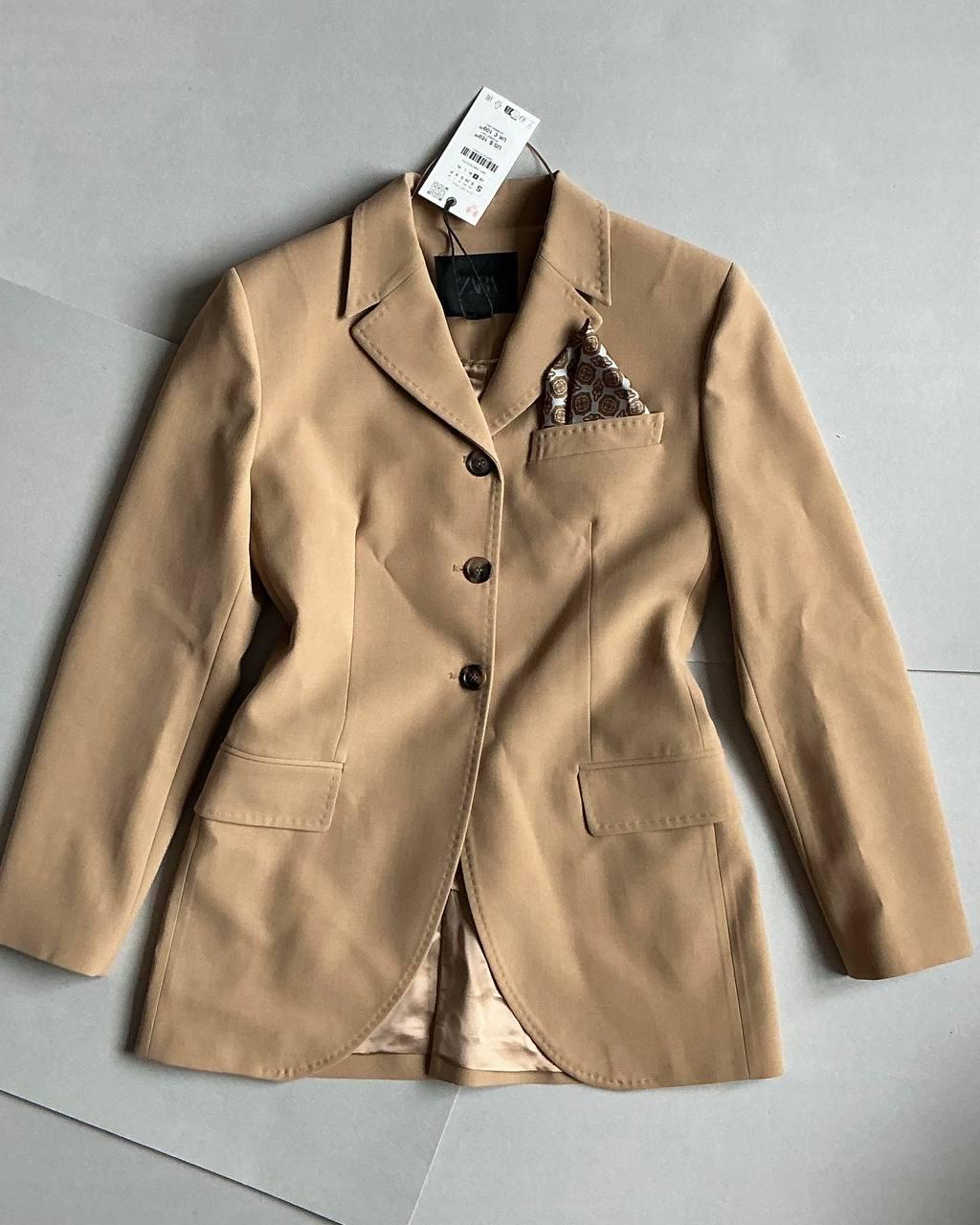Beige jacket with lapels and four buttons, patterned pocket square. Hanging tag visible against a grey background.